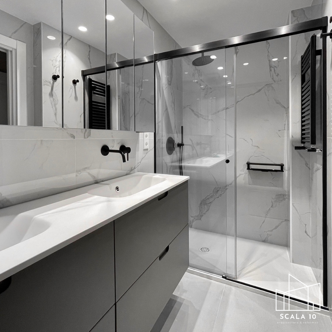 Baño moderno con paredes de mármol, lavabo de encimera blanca con grifería negra, espejo grande, ducha con puertas de vidrio, ducha de lluvia, toallero negro, y detalles negros en la decoración.
