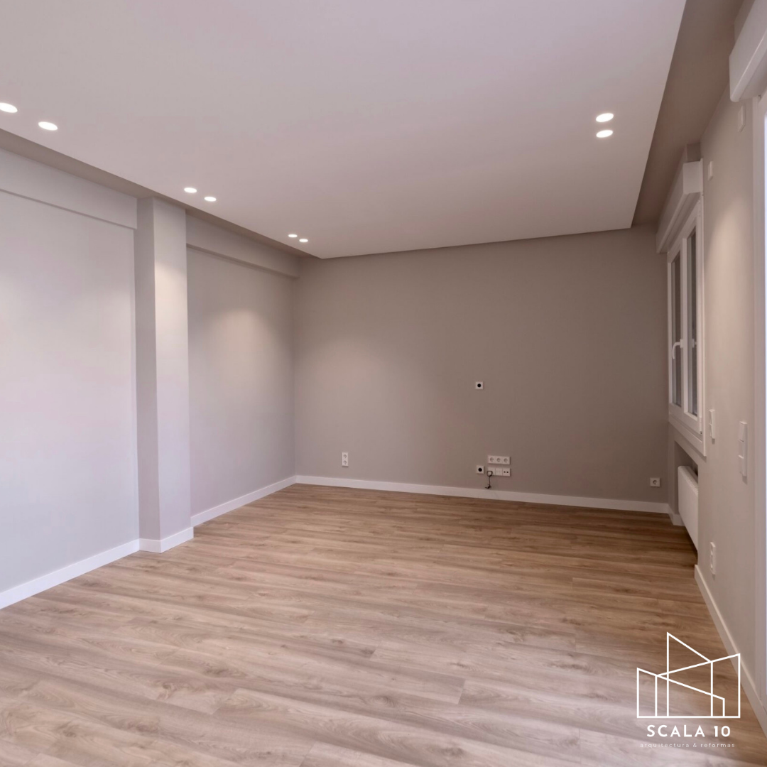 Sala vacía con paredes de color gris claro, suelo de madera clara, ventanas blancas y varias tomas eléctricas en la pared.