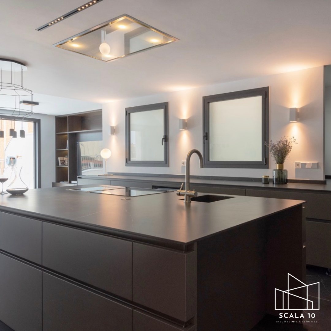 Cocina moderna con isla central gris, ventanas con marco gris, iluminación empotrada en el techo y paredes con apliques. Decoración minimalista con plantas y utensilios de cocina simples.