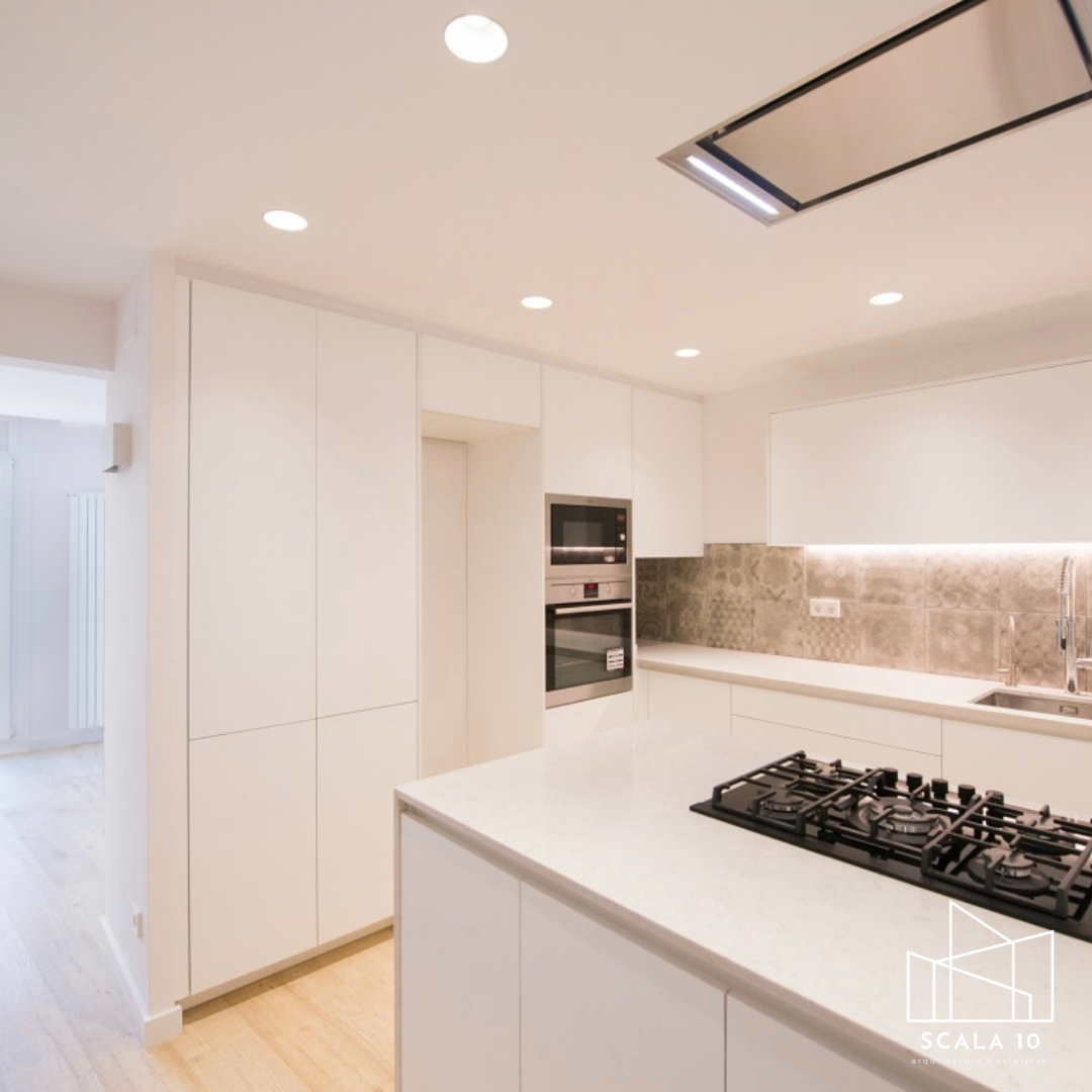 Cocina moderna con gabinetes blancos, encimera de granito, estufa de gas negra, horno empotrado y microondas, pared de azulejos decorados, iluminación empotrada y una ventana con persiana.