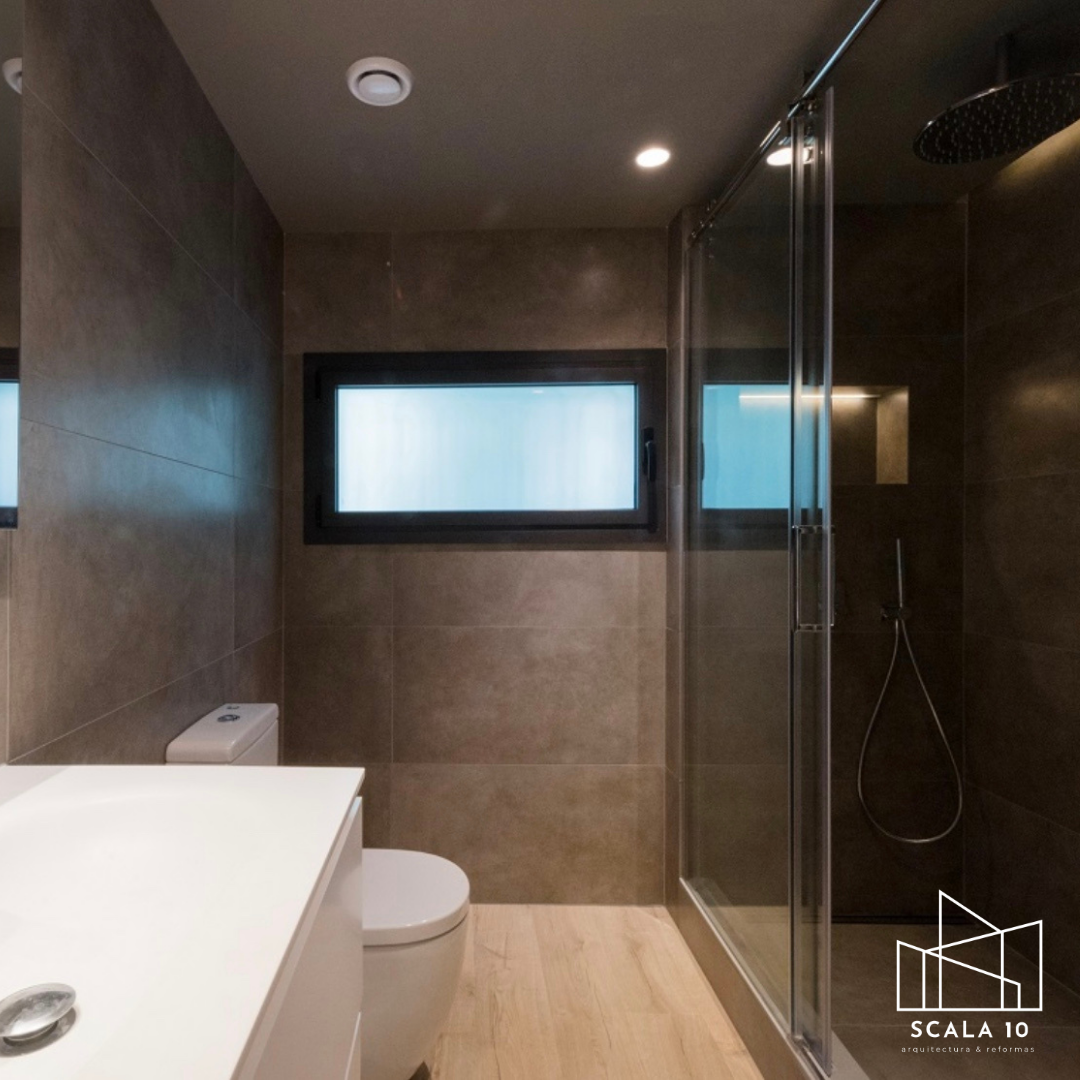 Baño moderno con paredes de piedra en tonos oscuros, ventana rectangular, ducha con mampara de cristal, inodoro y lavabo en blanco con encimera minimalista.