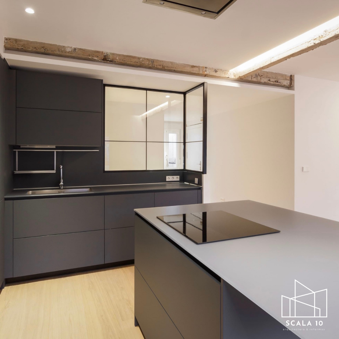 Cocina moderna con gabinetes grises y superficie de encimera blanca, isla central con estufa eléctrica, fregadero y una pared con ventanas de cristal divididas en cuatro paneles. Piso de madera y techo con iluminación empotrada.