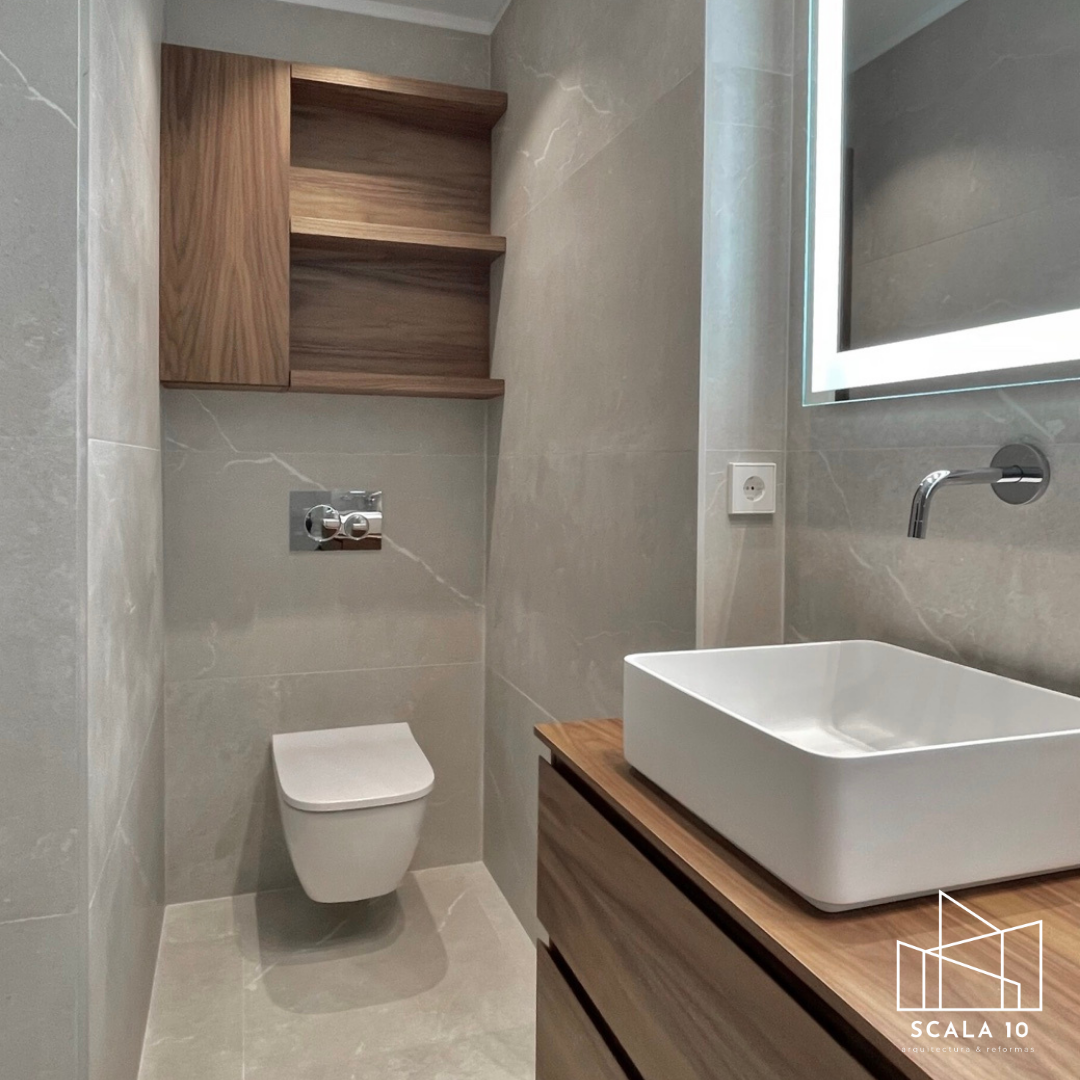 Baño moderno con lavabo de caja rectangular sobre mueble de madera, inodoro flotante, pared con mueble de madera y espejo con luz, paredes y suelo de mármol beige.