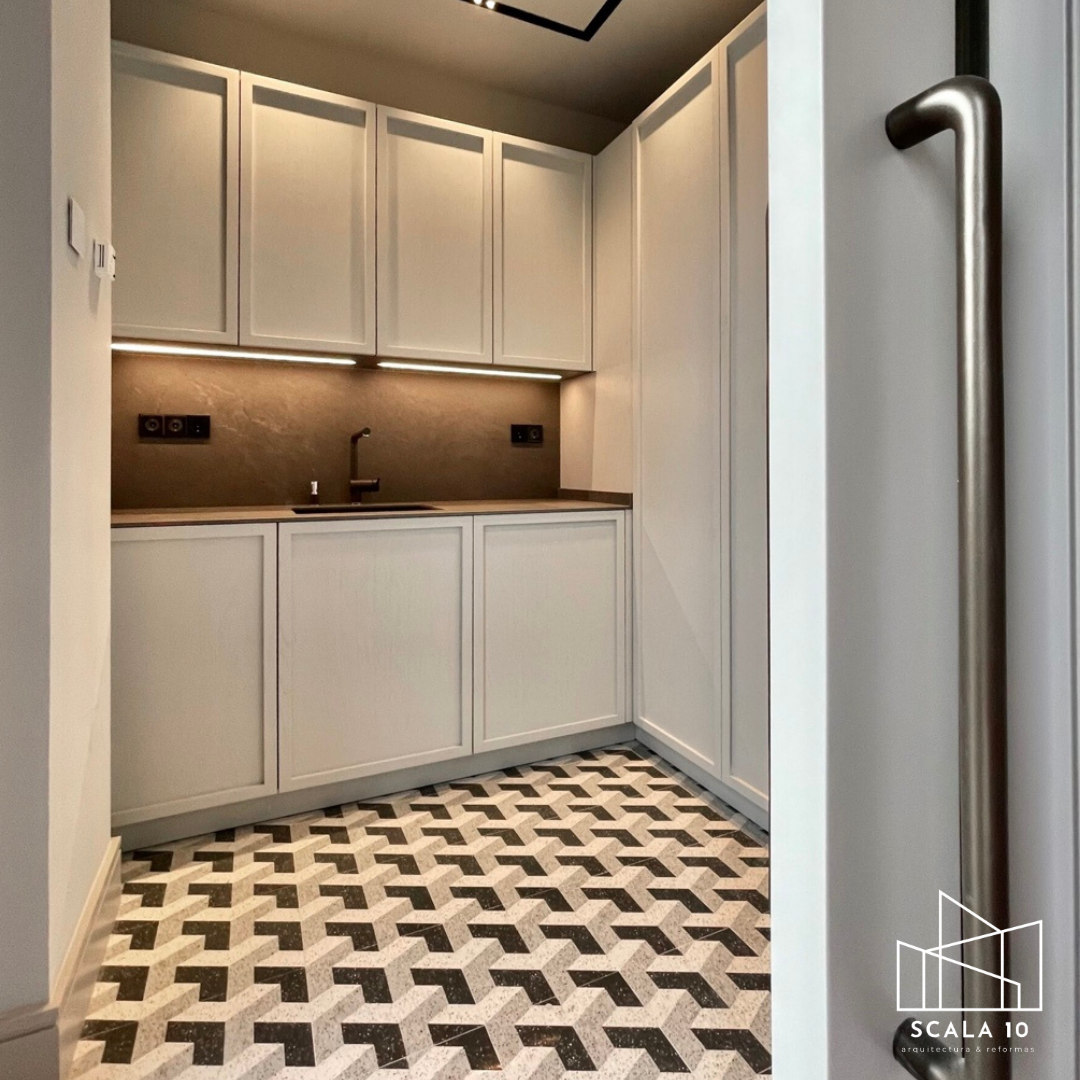 Pequeña cocina moderna con gabinetes blancos, encimera oscura, fregadero y grifo negro, iluminación debajo de los gabinetes, y piso con patrón geométrico en tonos grises y negros.