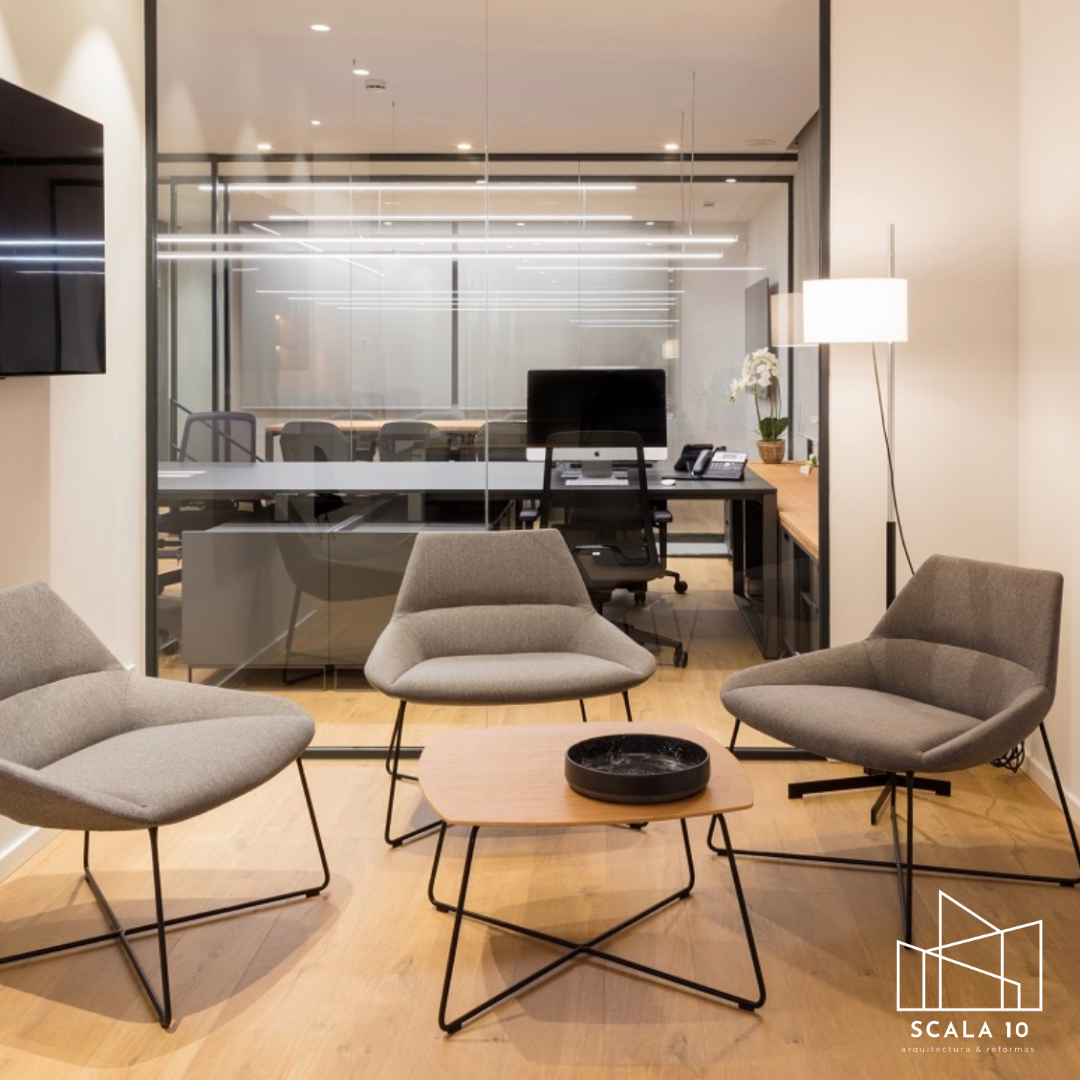 Sala de espera con tres sillones grises y una mesa de centro de madera con bandeja negra. Detrás, una oficina con escritorio, silla y computadora, separada por una pared de vidrio.