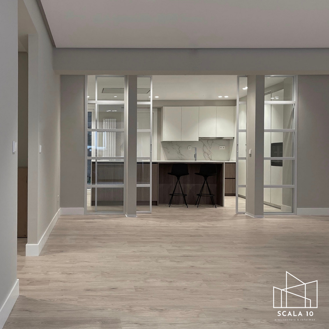 Interior de una sala moderna con suelo de madera clara, paredes blancas y un área de cocina al fondo separado por un divisor de vidrio con perfiles blancos. La cocina tiene gabinetes blancos, encimera de mármol y una isla con dos sillas negras.