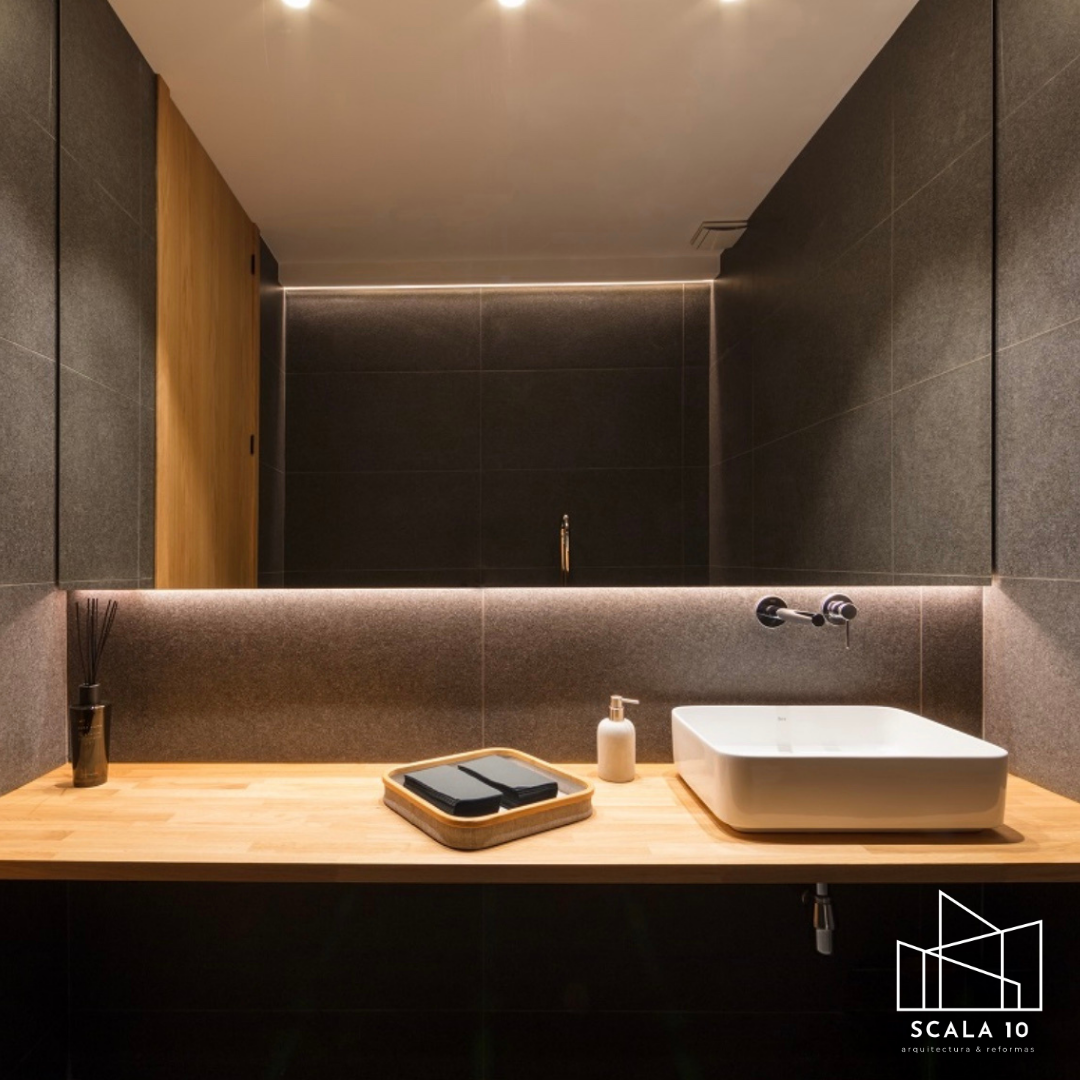 Lavabo rectangular blanco en un mostrador de madera, con espejo grande detrás, paredes de azulejo gris oscuro, dispensador de jabón y un bote con varillas aromáticas.