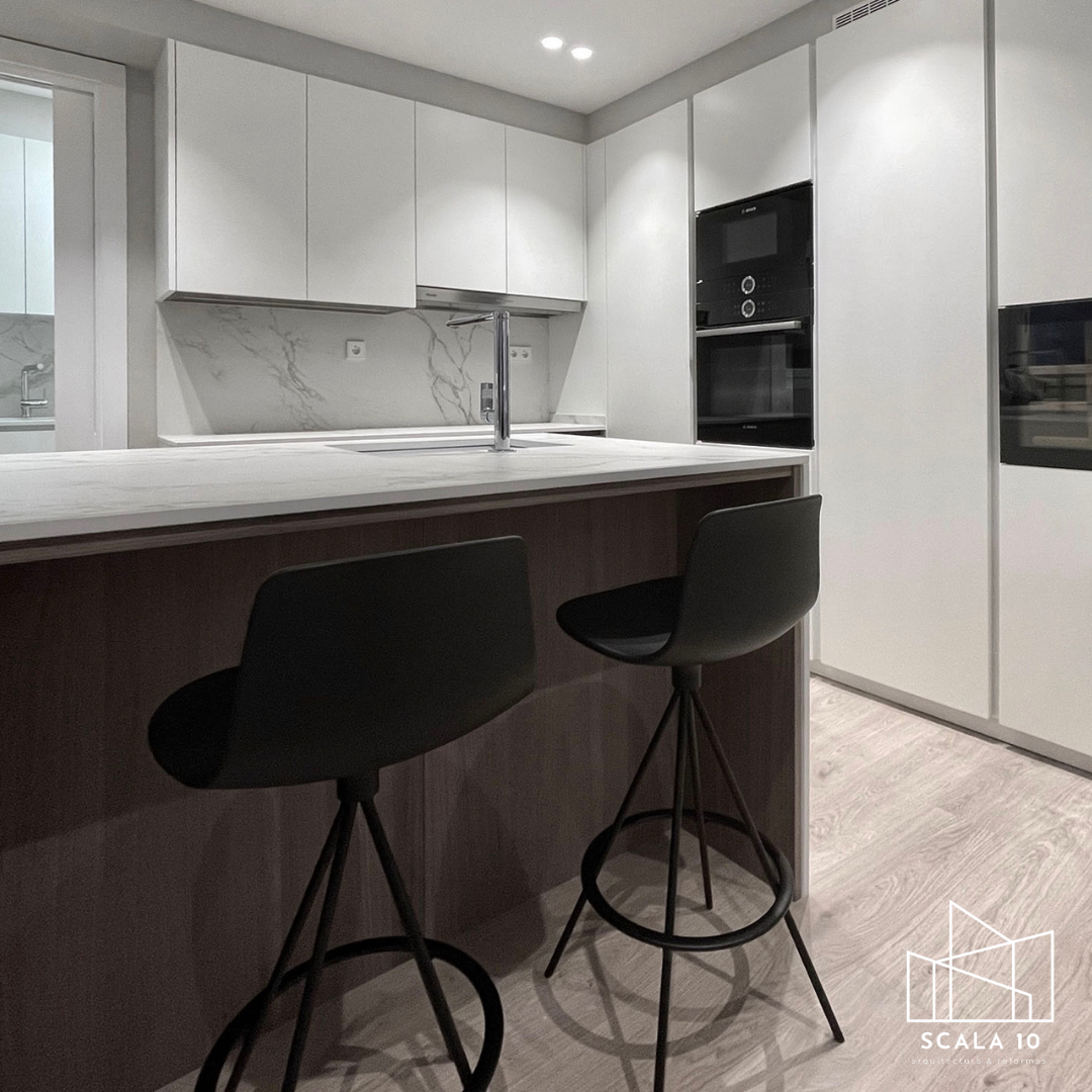 Cocina moderna con encimera de mármol, gabinetes blancos, una isla con barandas oscuras, dos sillas negras y electrodomésticos integrados de color negro.