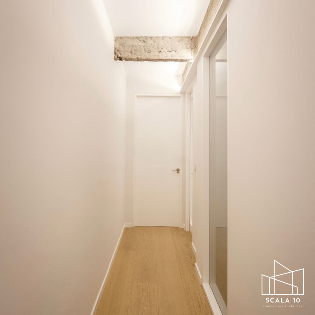 Pasillo interior con paredes blancas, suelo de madera clara, y cerca del techo una viga de cemento expuesta. Al fondo una puerta blanca. A la derecha hay una puerta de elevador con paredes blancas y piso de madera.