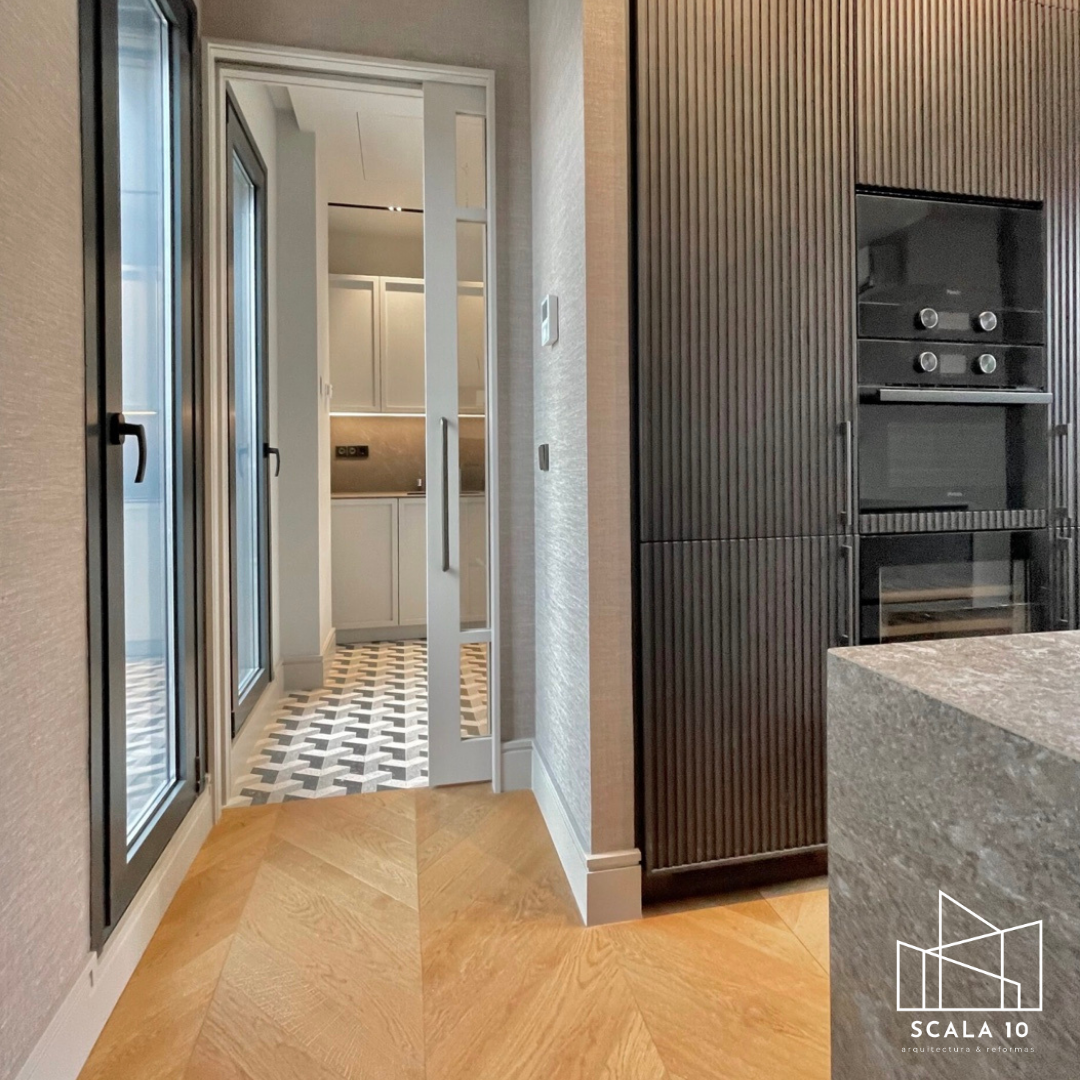 Vista interior de una cocina moderna en tonos neutros, con puertas de vidrio en el fondo y suelo de baldosas en patrón geométrico.