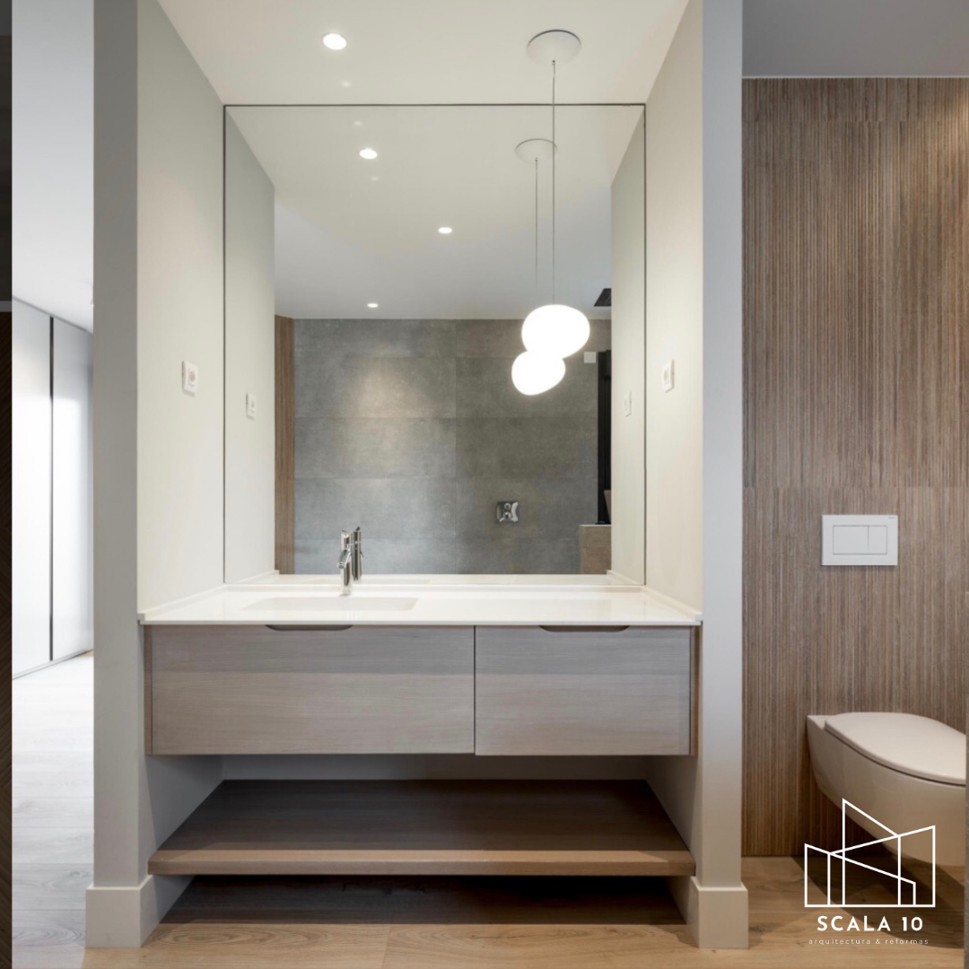 Espejo grande sobre lavabo de baño con espejo, paredes blancas y una pared de madera en el fondo, con una lámpara colgante de tres globos blancos.