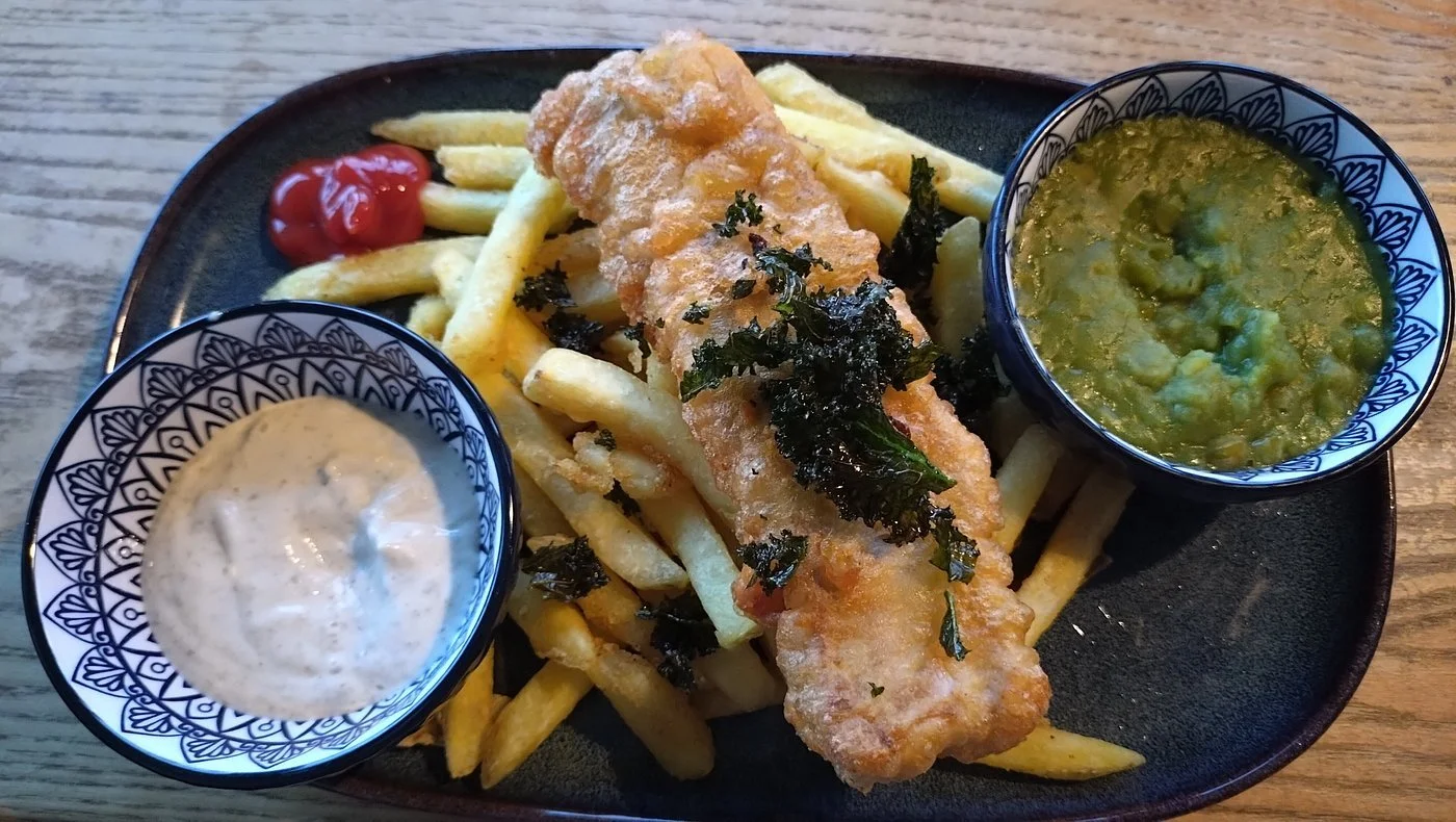Fried fish fillet on a bed of French fries with two bowls of dipping sauces, one creamy white and one green, on a black oval plate. Newhaven Pub & Restaurant