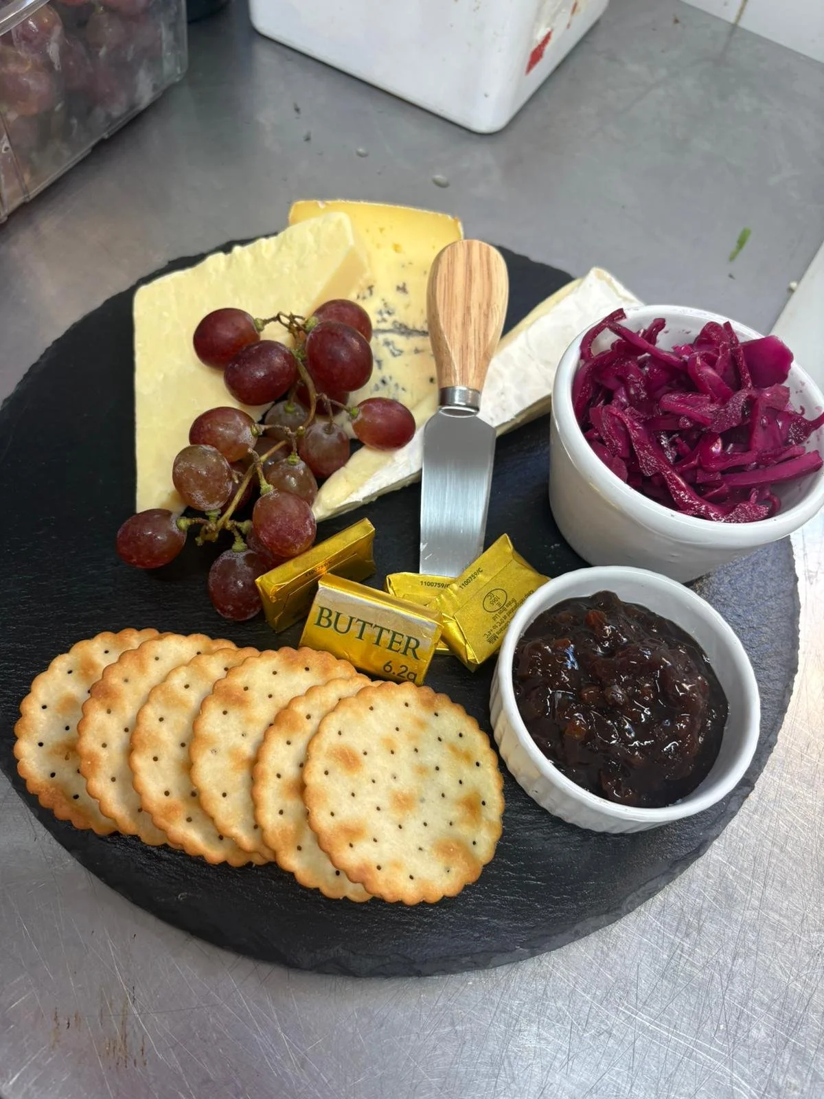 A cheese and cracker platter with grapes, butter, red onion slaw, blackberry jam, and sliced cheese on a round black slate board.