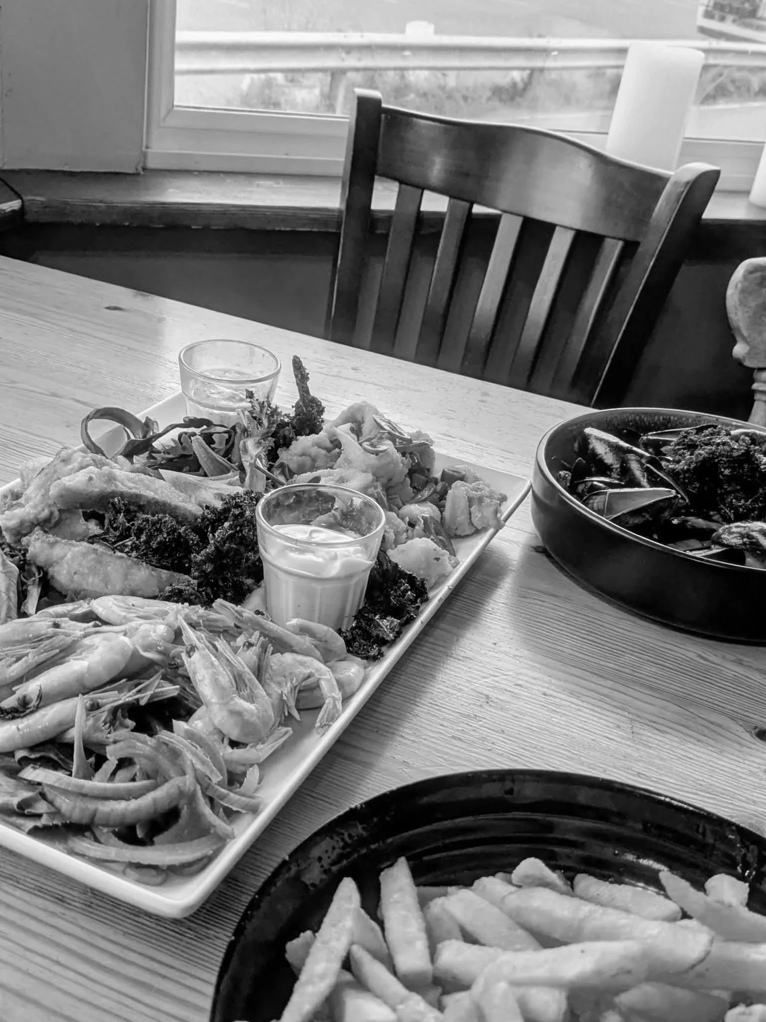 A table set with a large platter of fried seafood and vegetables, small glasses of sauce, a bowl of mussels, and a bowl of French fries, near a window with a wooden chair in the background. Newhaven Pub & Restaurant
