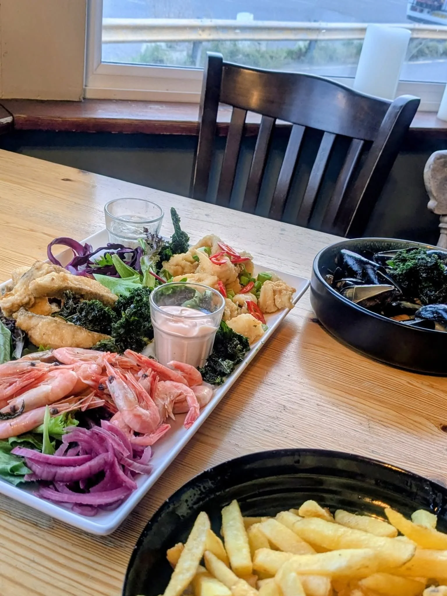 A meal featuring a large plate with shrimp, fried fish fillets, and an assortment of vegetables including purple onions, cherry tomatoes, and broccoli, along with two small cups of dipping sauce. Also, a black bowl with steamed mussels and a black di