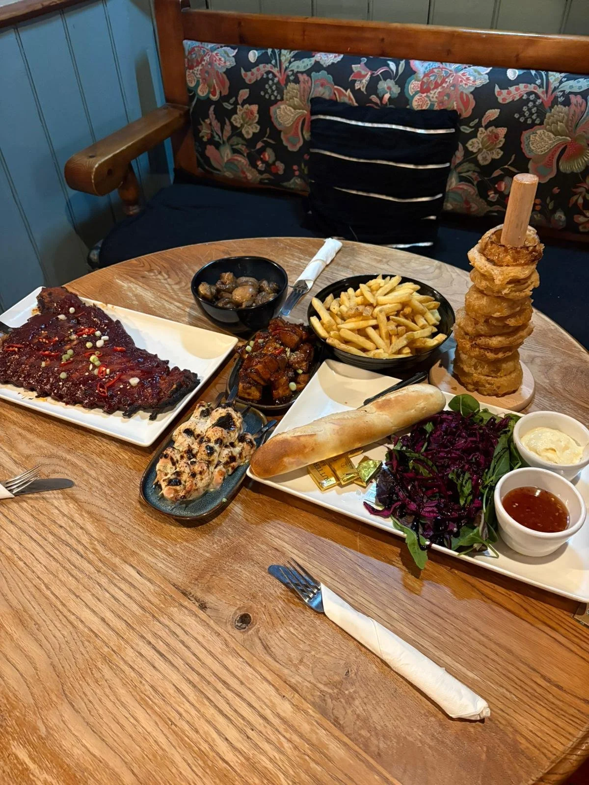 A wooden table with various dishes including French fries, onion rings, ribs, grilled chicken skewers, a bread roll, beef brisket, purple cabbage salad, and dipping sauces in white bowls, set in a restaurant with a blue wall and floral patterned boot