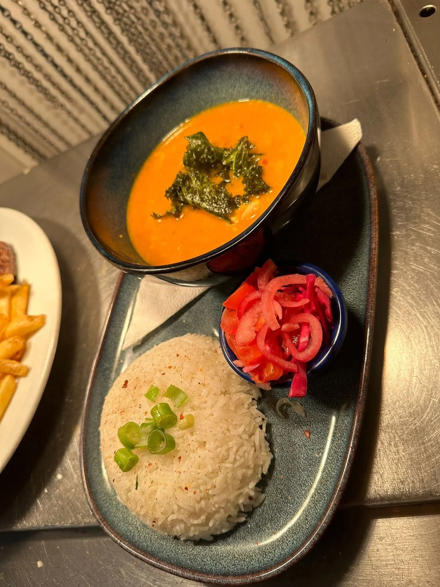 A plate with a serving of white rice topped with chopped green onions, a bowl of orange curry garnished with greens, and a small bowl of pickled red onions.