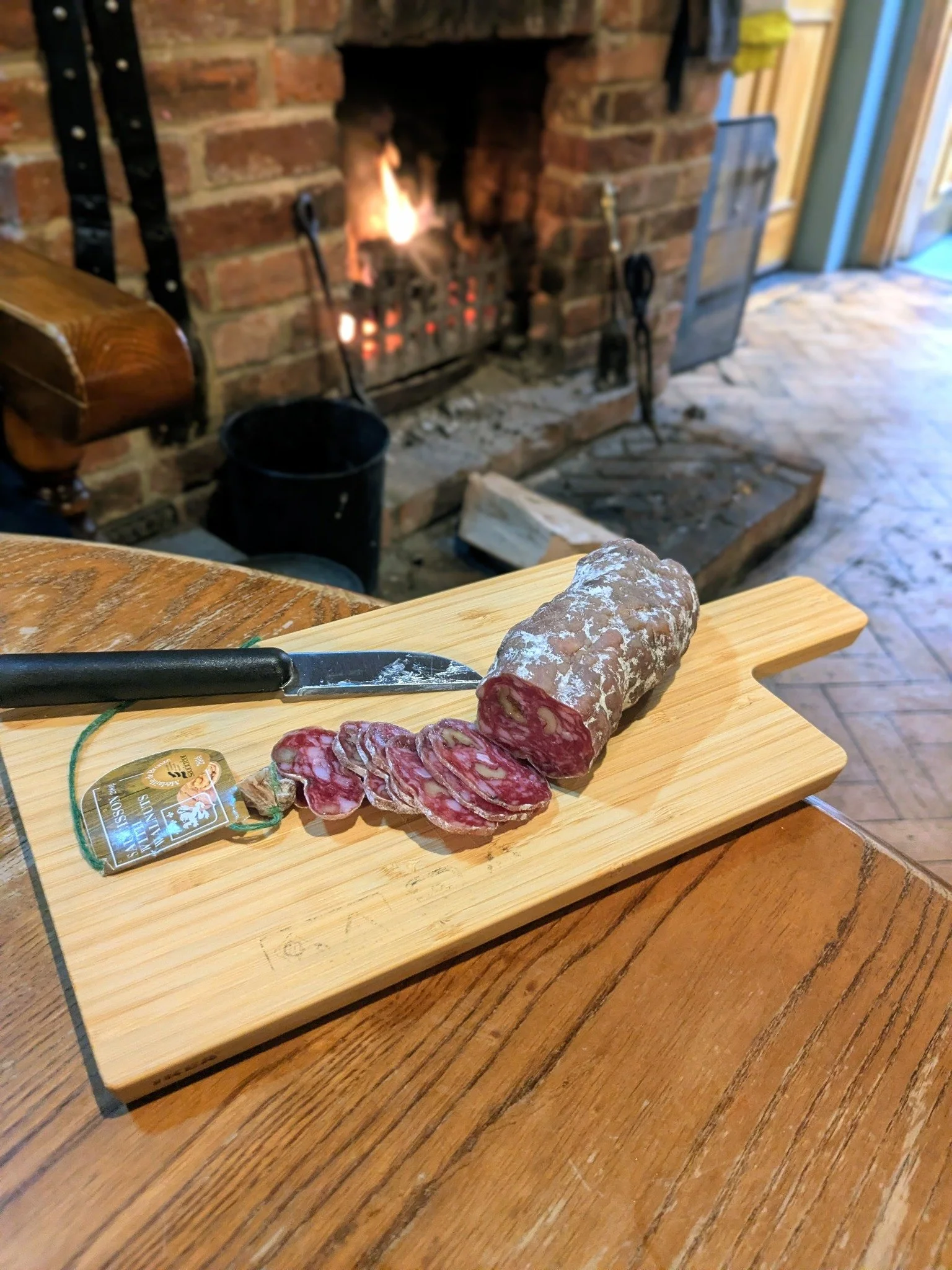 Sliced salami on a wooden cutting board with a knife, in front of a brick fireplace with a fire burning.