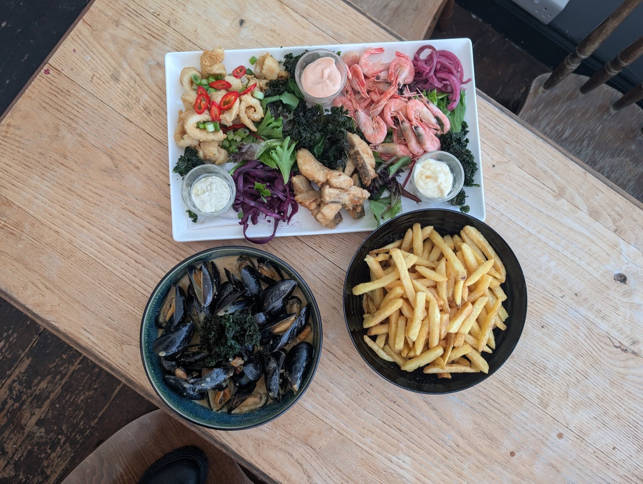 A table with three dishes: a rectangular platter of seafood salad with shrimp, calamari, vegetables, and sauces; a bowl of French fries; a bowl of cooked mussels garnished with herbs.