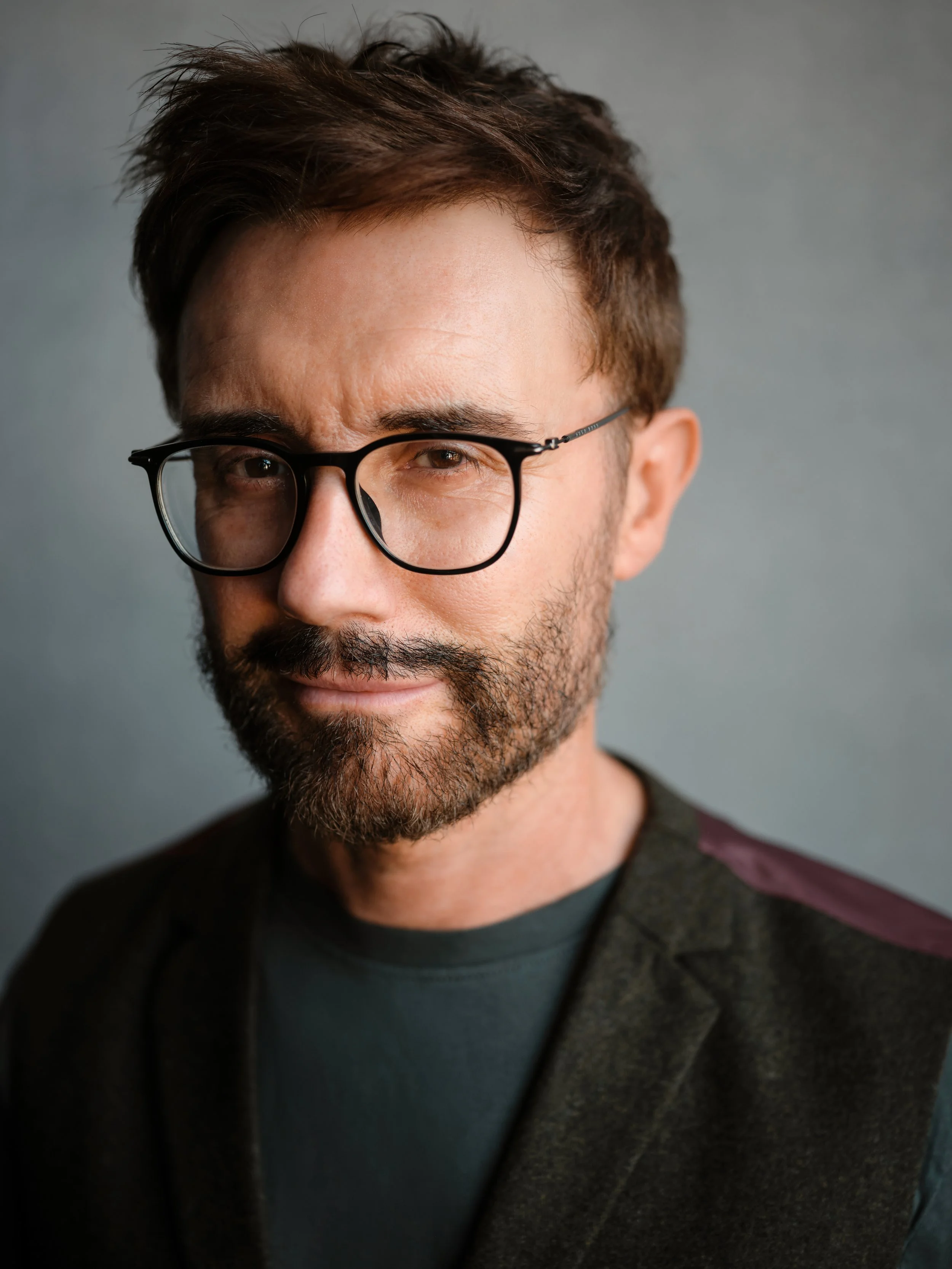 Close-up portrait of a man with brown hair, beard, and glasses, smiling slightly against a plain gray background.