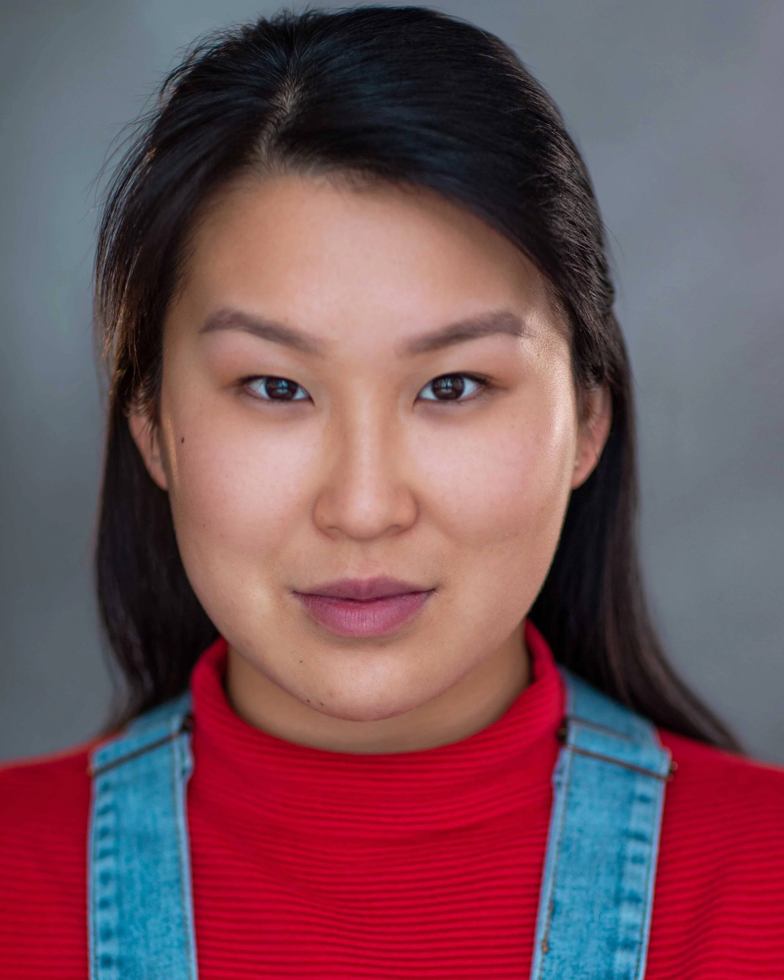 Close-up of a woman with dark hair, wearing a red top and denim overalls, looking directly at the camera against a plain background.