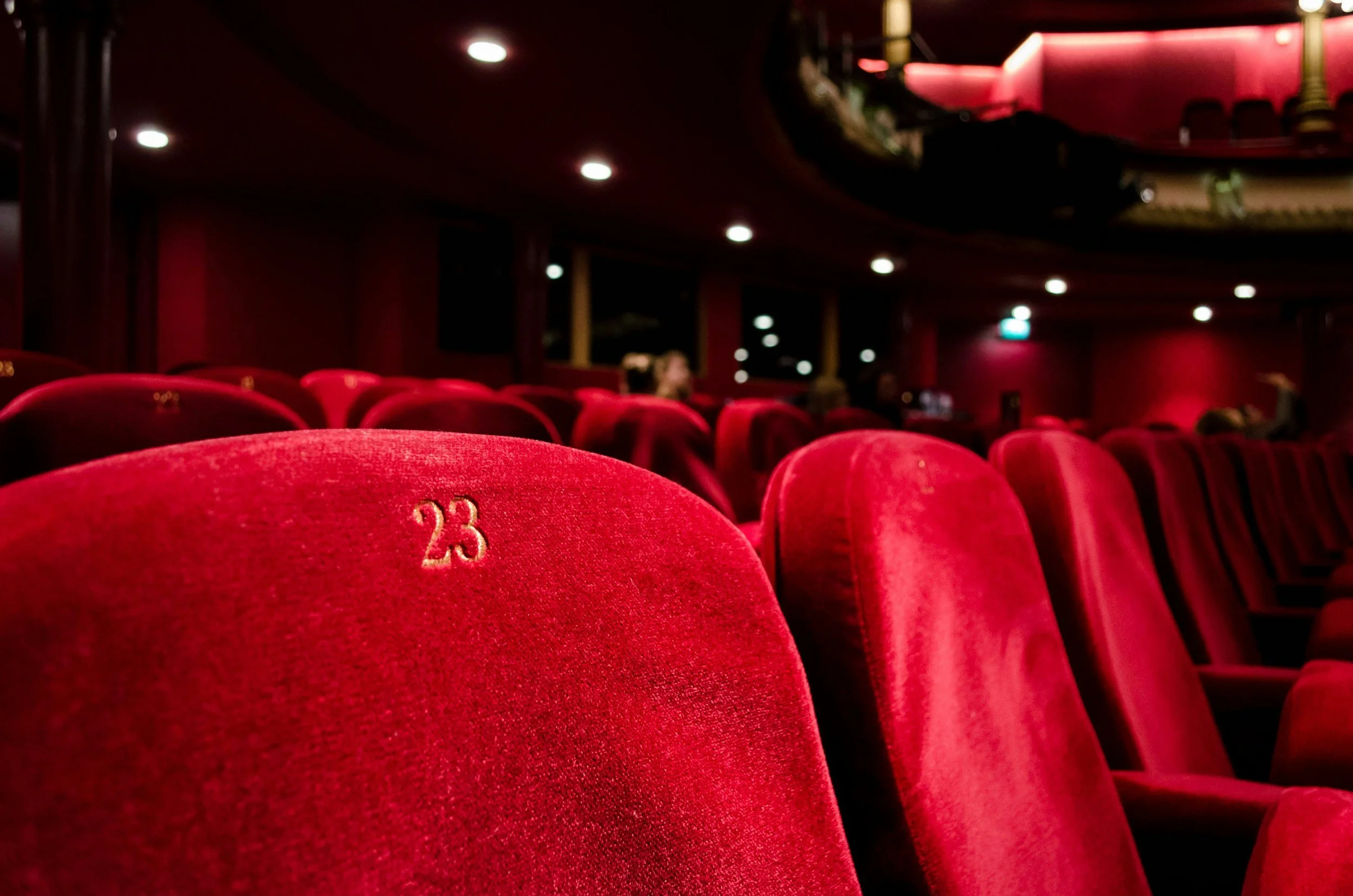 Red theater seats with the number 23 embroidered on one seat in an auditorium or theater, dim lighting, and a curved balcony above.