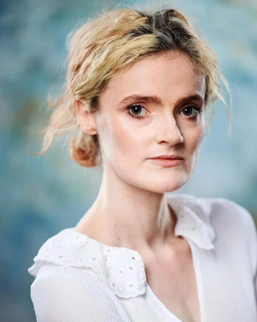 Close-up portrait of Nim McQuin, a woman with blonde, wavy hair, wearing a white blouse with eyelet details on the collar, against a blurred blue-green background.