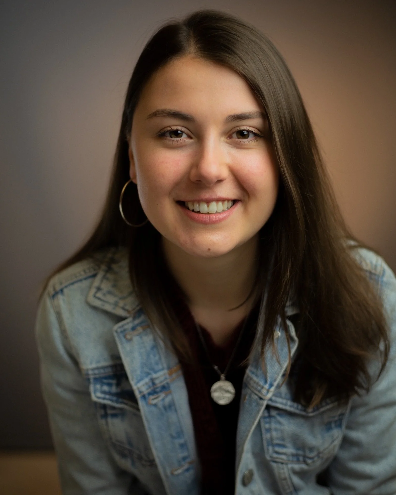 Abbie Atkinson, the host of Don't Let Dave Win podcast, a young woman with long brown hair, wearing hoop earrings and a denim jacket, smiling at the camera against a plain background.