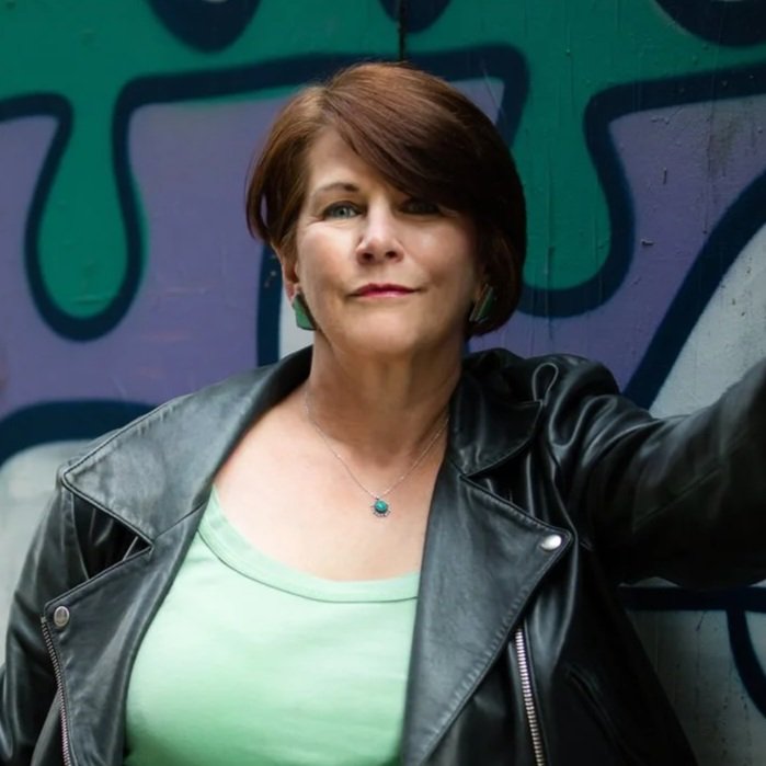 Stacia Keogh, the host of Finding Your Voice podcast, a woman with short reddish-brown hair taking a selfie in front of a colorful graffiti wall, wearing a black leather jacket and a light green shirt.
