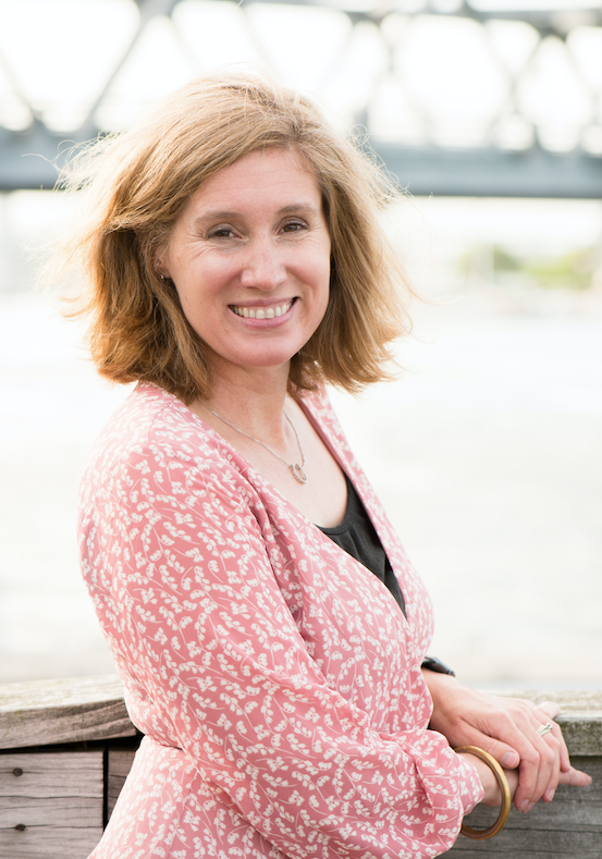 A smiling woman with shoulder-length, wavy, blonde hair stands outdoors near a wooden railing with a bridge over water in the background.