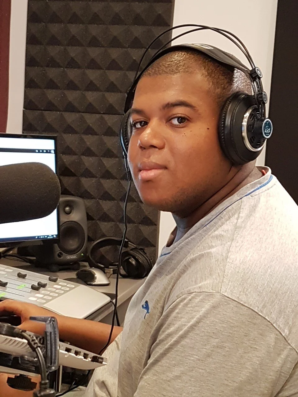 A young man wearing large over-ear headphones sitting at a radio station or recording studio with soundproof padding on the wall, a microphone, speakers, and audio equipment.