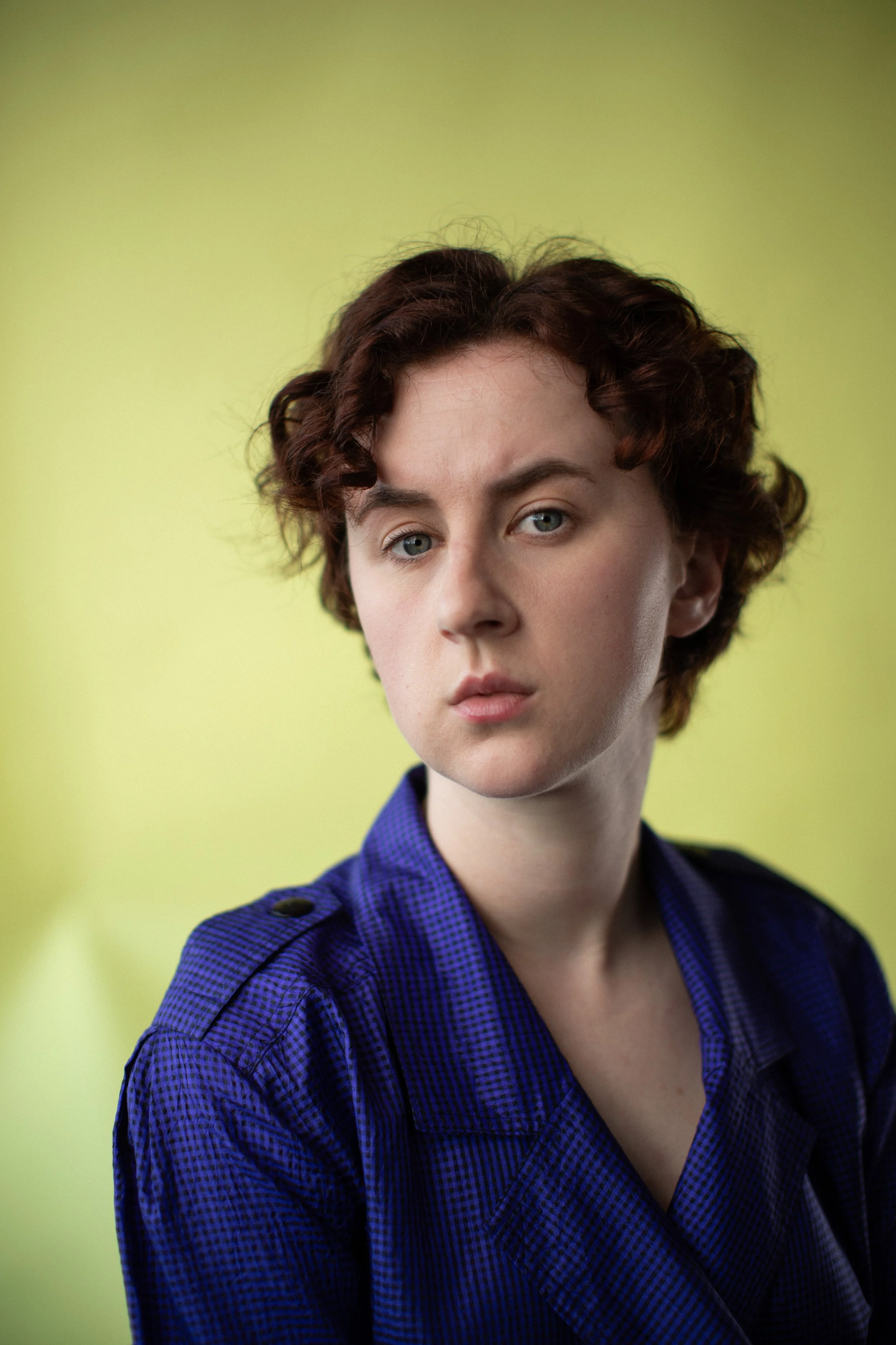 Portrait of a young woman with short, curly red hair and blue eyes, wearing a dark blue patterned shirt, against a light yellow background.