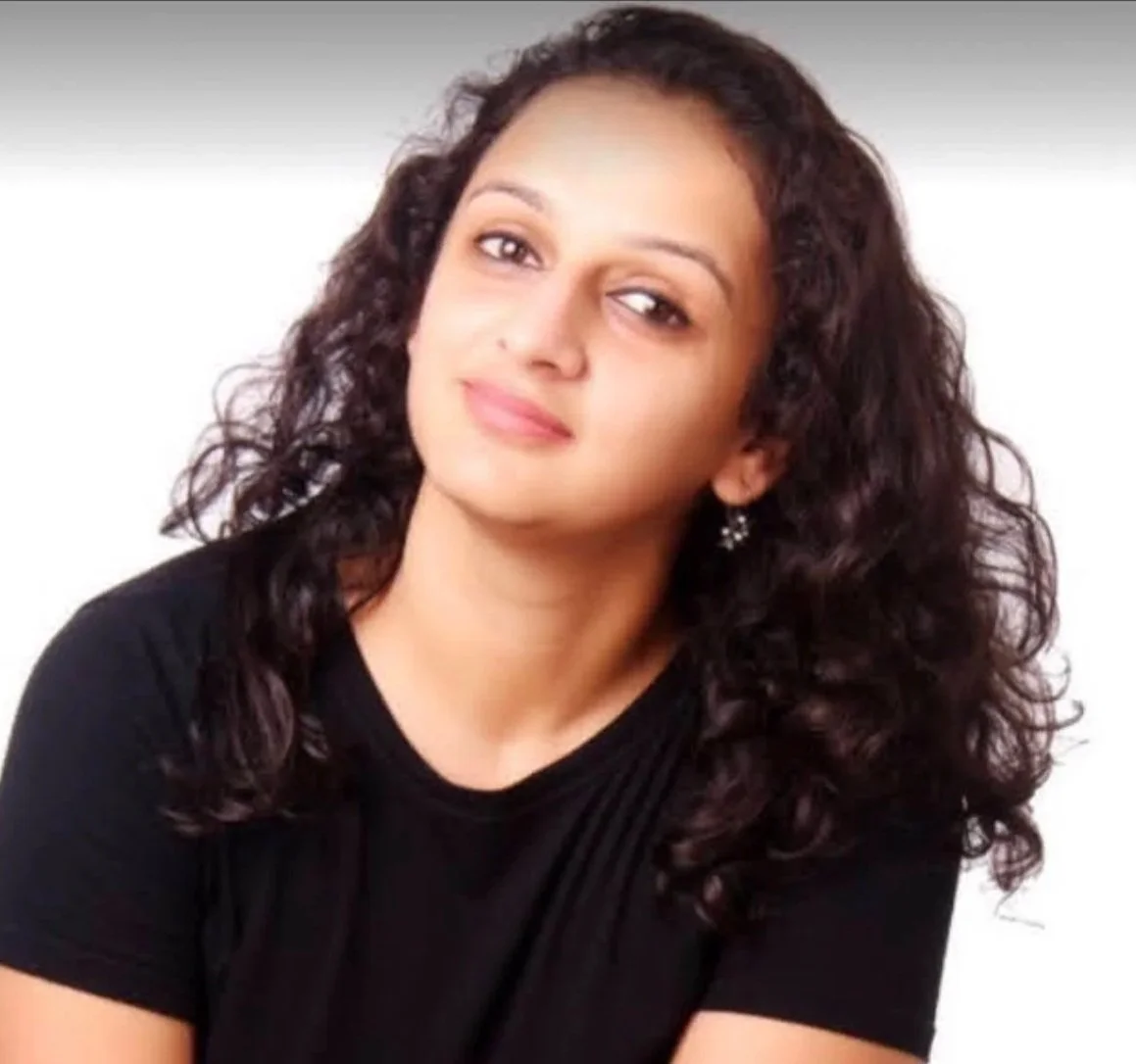 A woman with curly dark hair, wearing a black top and earrings, looking at the camera with a slight smile against a light background.