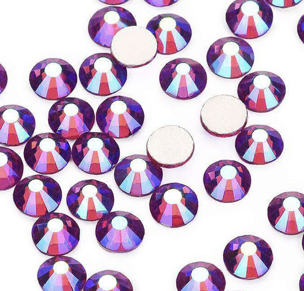 Close-up of purple rhinestones with reflective surfaces scattered on a white background, showing some rhinestones with smooth backing.