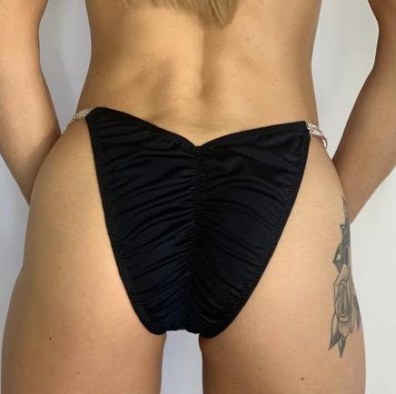 Close-up of a person wearing black ruched bikini bottoms, showing their lower abdomen and tattoo on their right thigh.