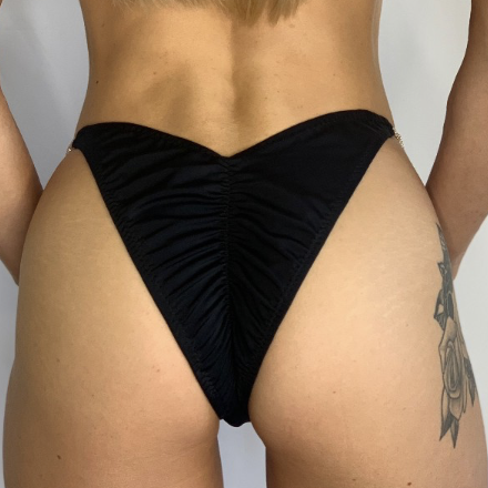 Close-up of a woman's lower abdomen and hips wearing black ruched bikini bottoms, with a tattoo visible on her right thigh.