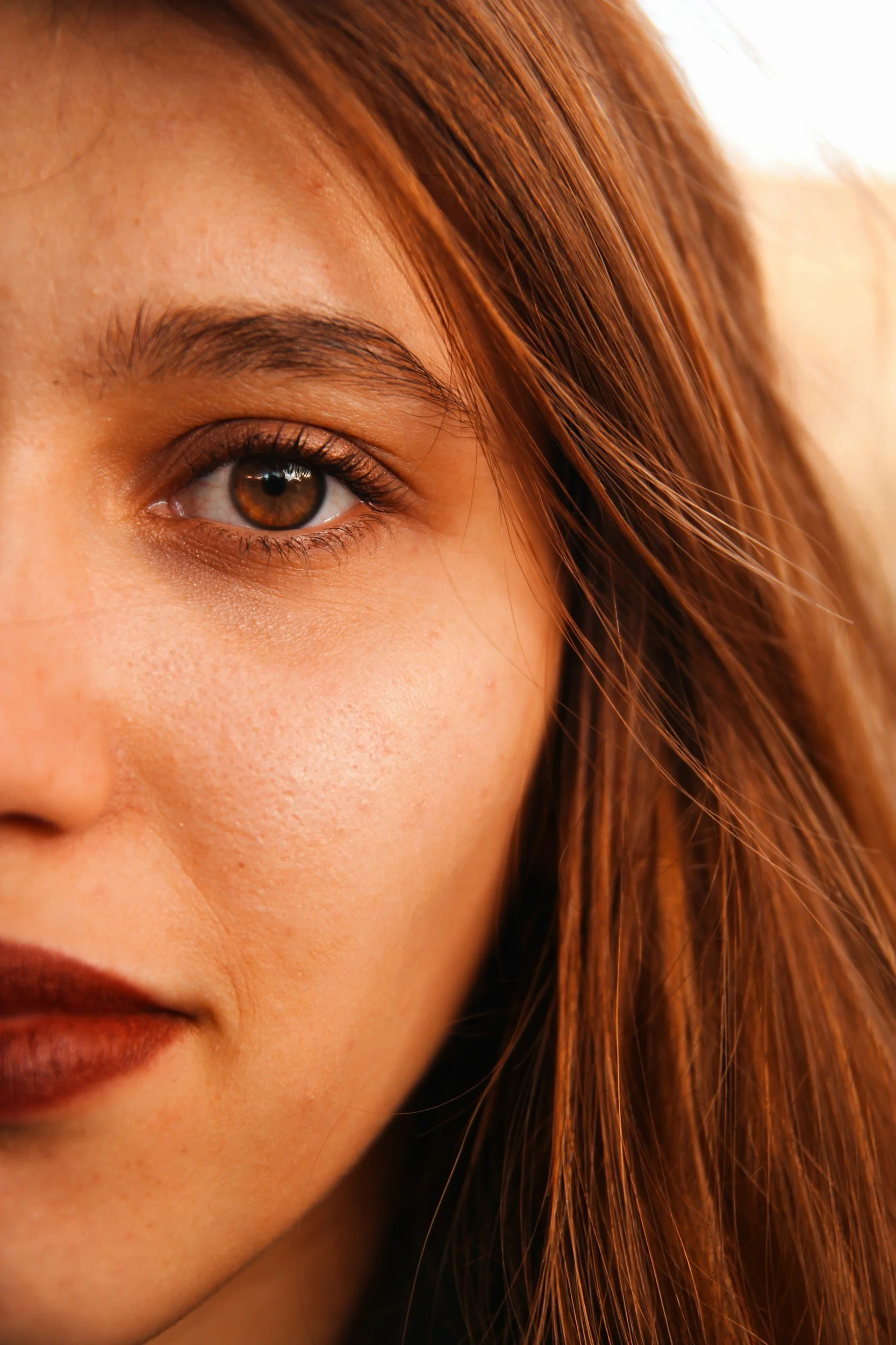 Close-up of a woman's face, showing her eye, eyebrow, cheek, and part of her lips, with long, wavy auburn hair.
