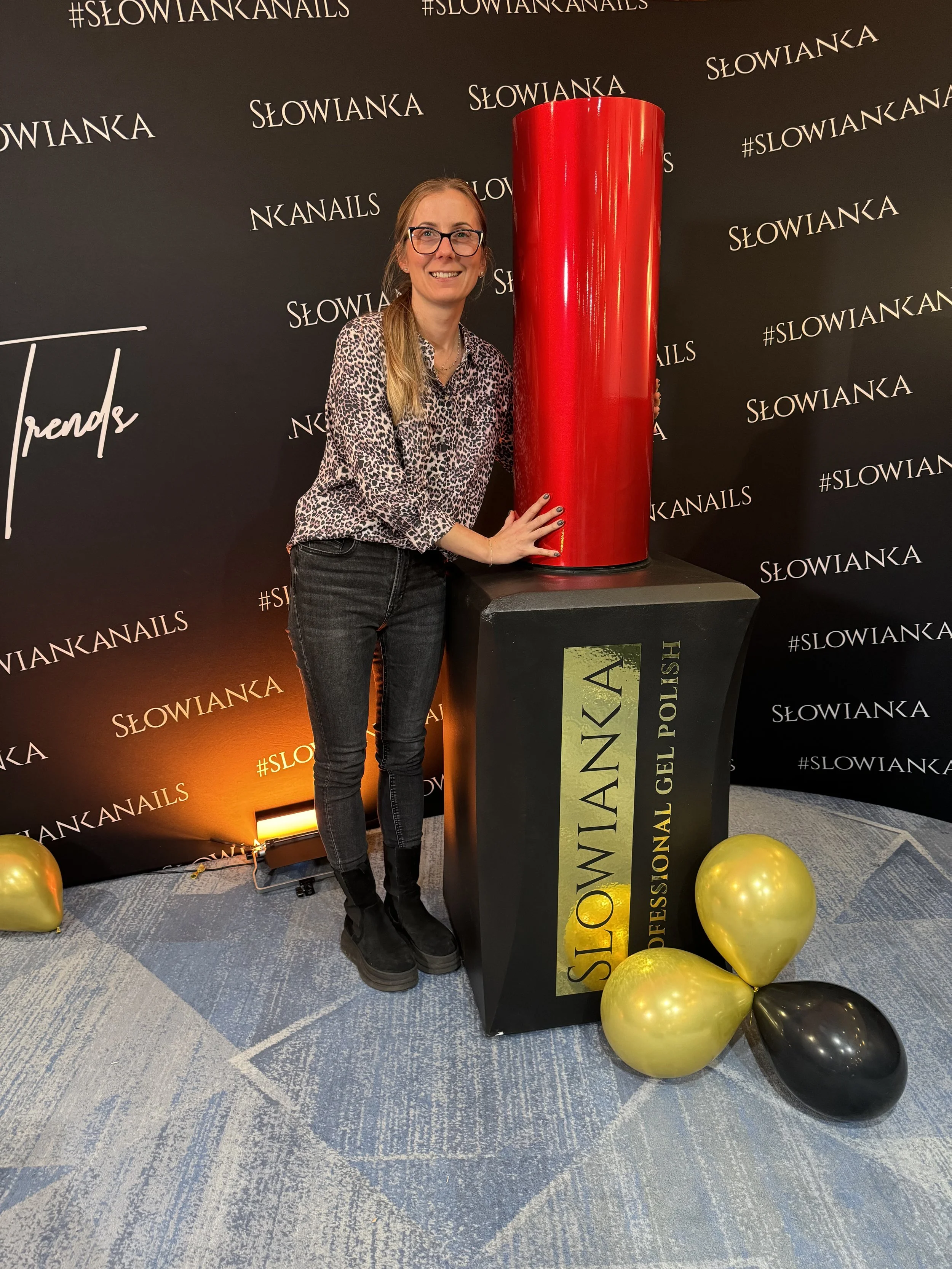 A woman standing next to a large red cosmetic product display, with SLOWIANKA branding on a black backdrop and balloons on the floor.