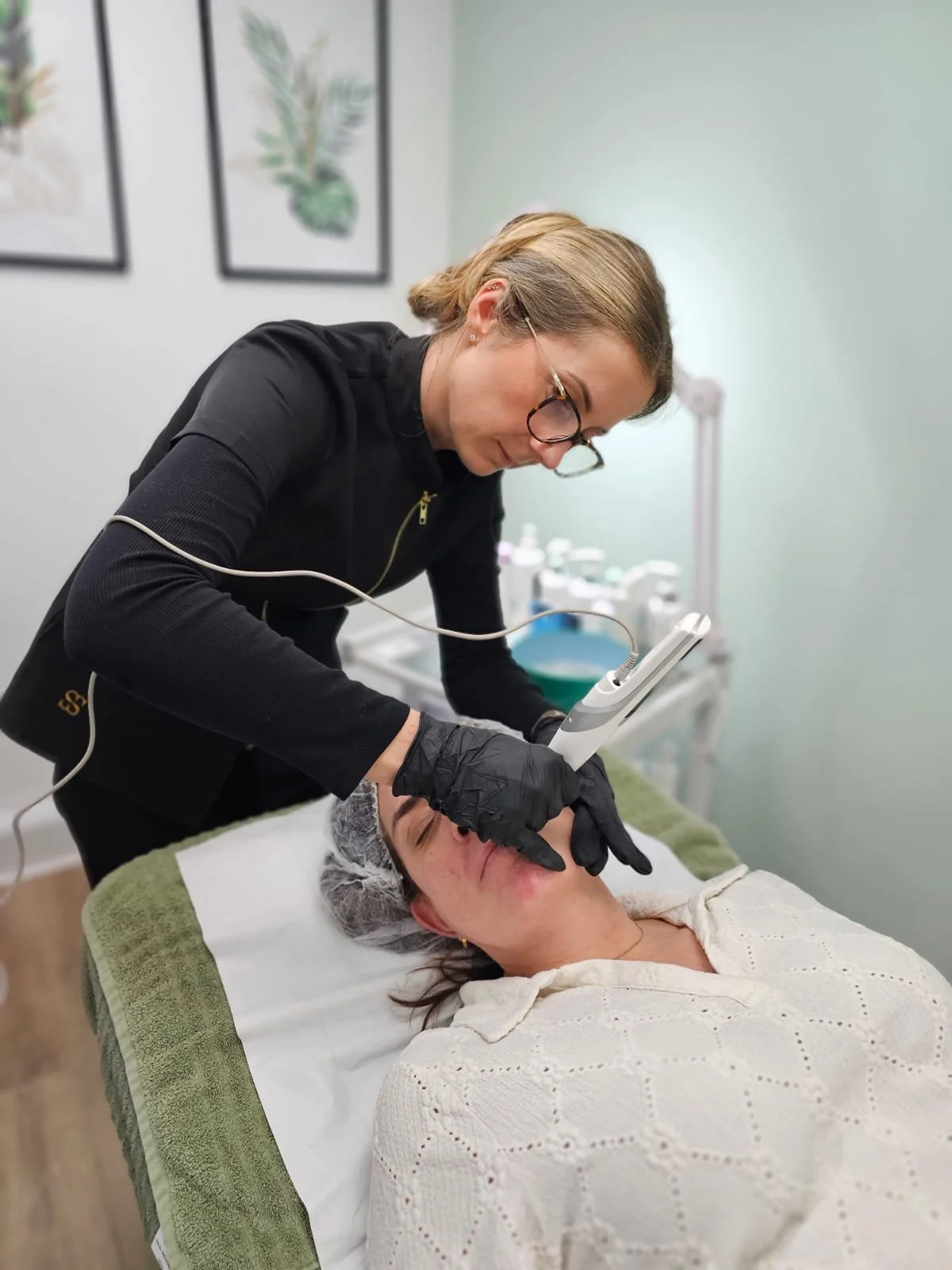 A woman receives a facial treatment from a professional in a clinic or spa setting, lying on a treatment bed covered with a white towel, with her head wrapped in a disposable shower cap. A facial is being performed for problematic skin.