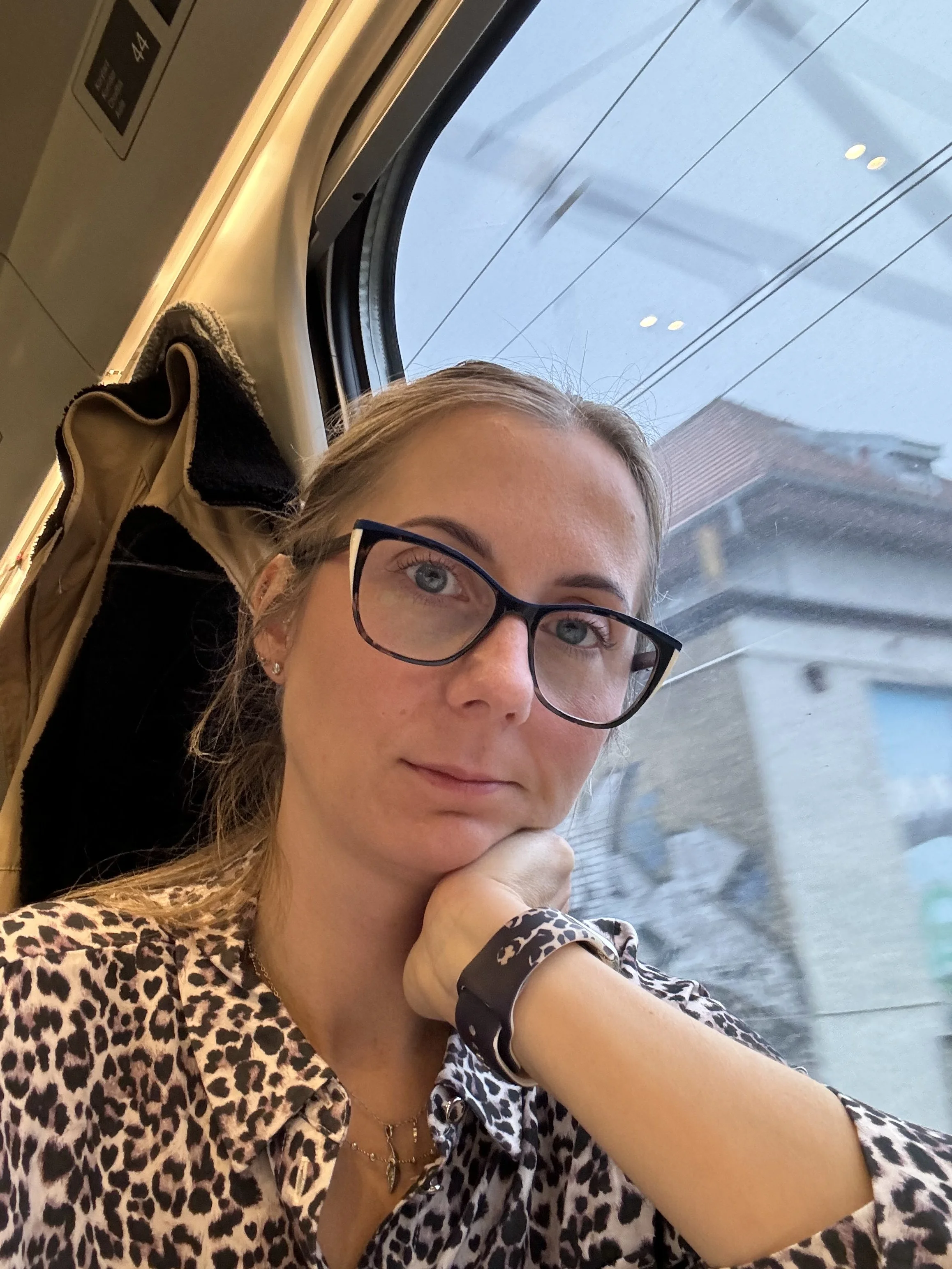 A young woman with glasses and leopard print clothing sitting on a train, resting her chin on her hand, looking at the camera with a building and power lines outside the window behind her.
