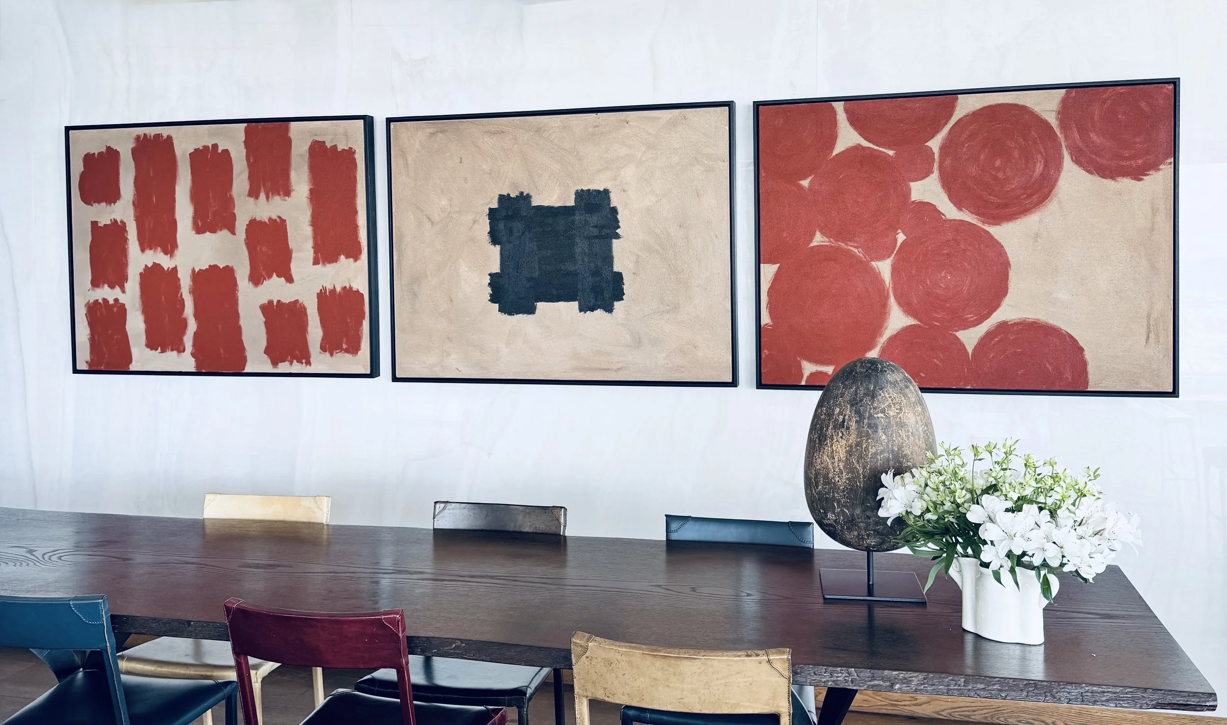 Dining room with a wooden table, colorful chairs, large abstract paintings on the wall, a decorative vase, and a floral arrangement.