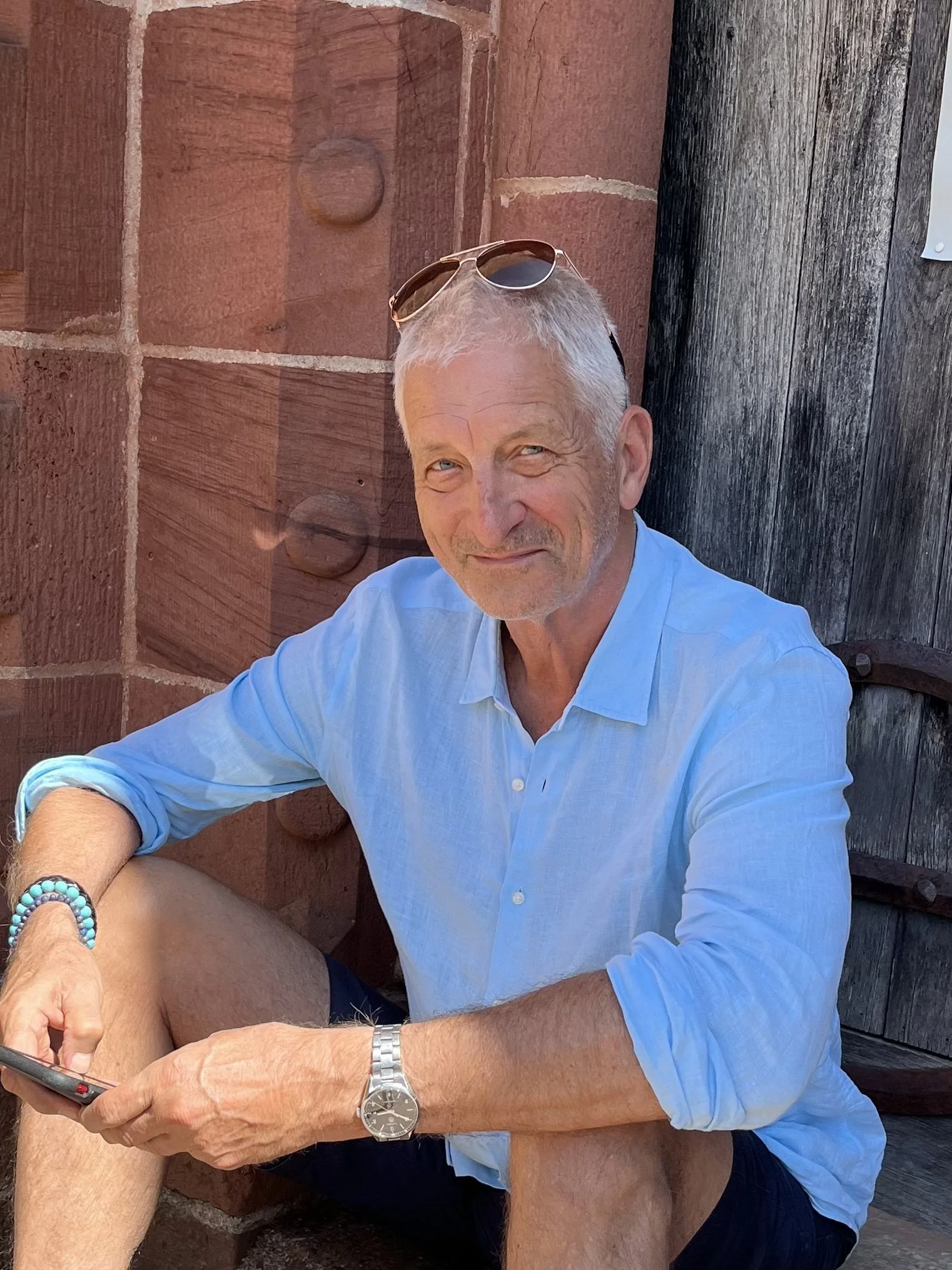 A smiling older man in a light blue shirt sitting outdoors near a brick wall and wooden door, with sunglasses resting on his head, holding a smartphone.