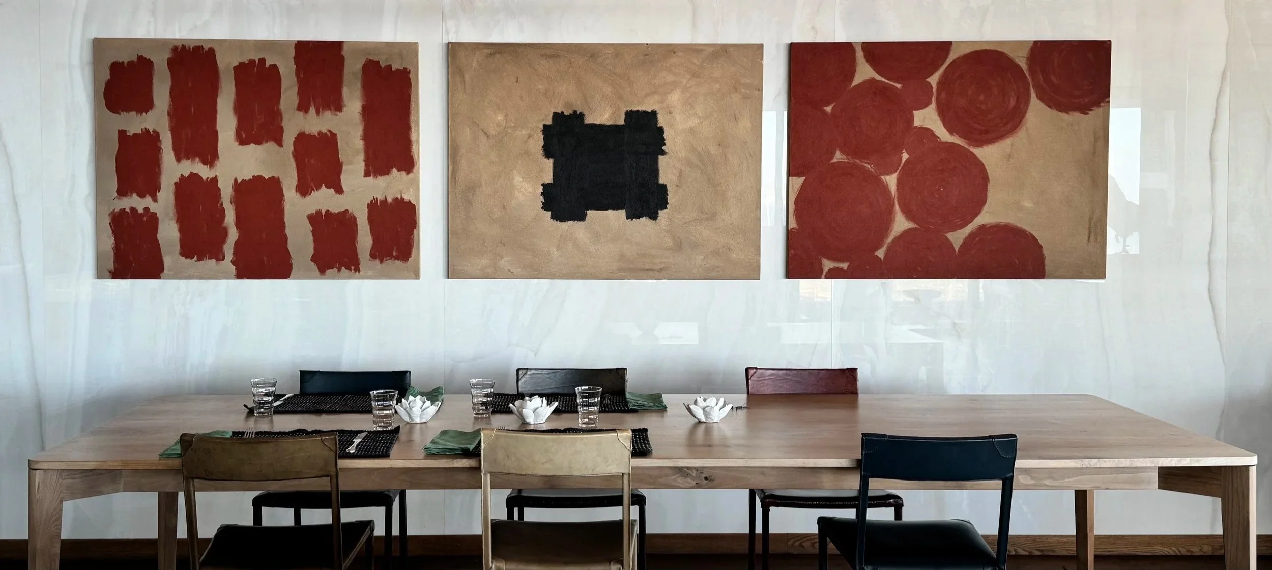 A dining table set with glassware, napkins, and decorative paper flowers, against a wall with three abstract paintings featuring red and black shapes.
