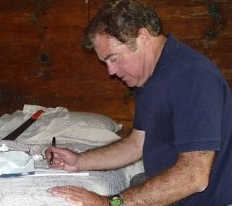 Author Carl Martin Johnson, pictured in a blue shirt writing at a desk in a room with wooden walls.