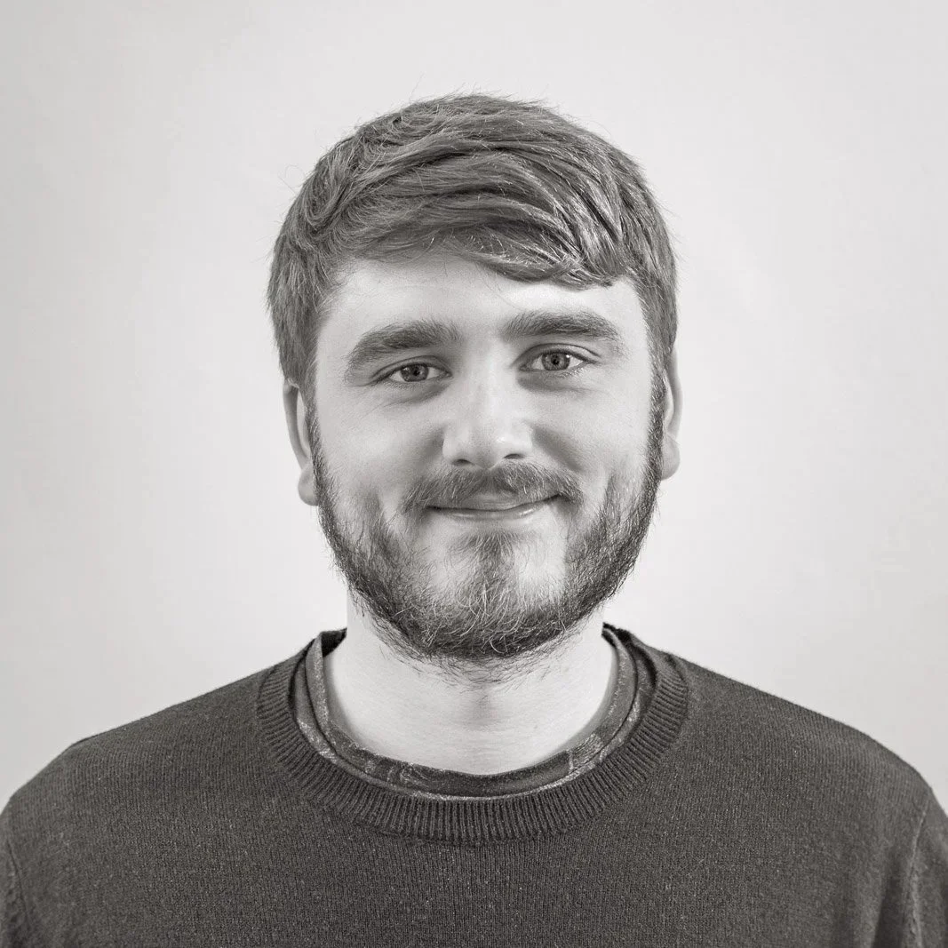 Black and white portrait of a young man with light-colored eyes, short hair, and a full beard, smiling and wearing a dark sweater, against a plain background.