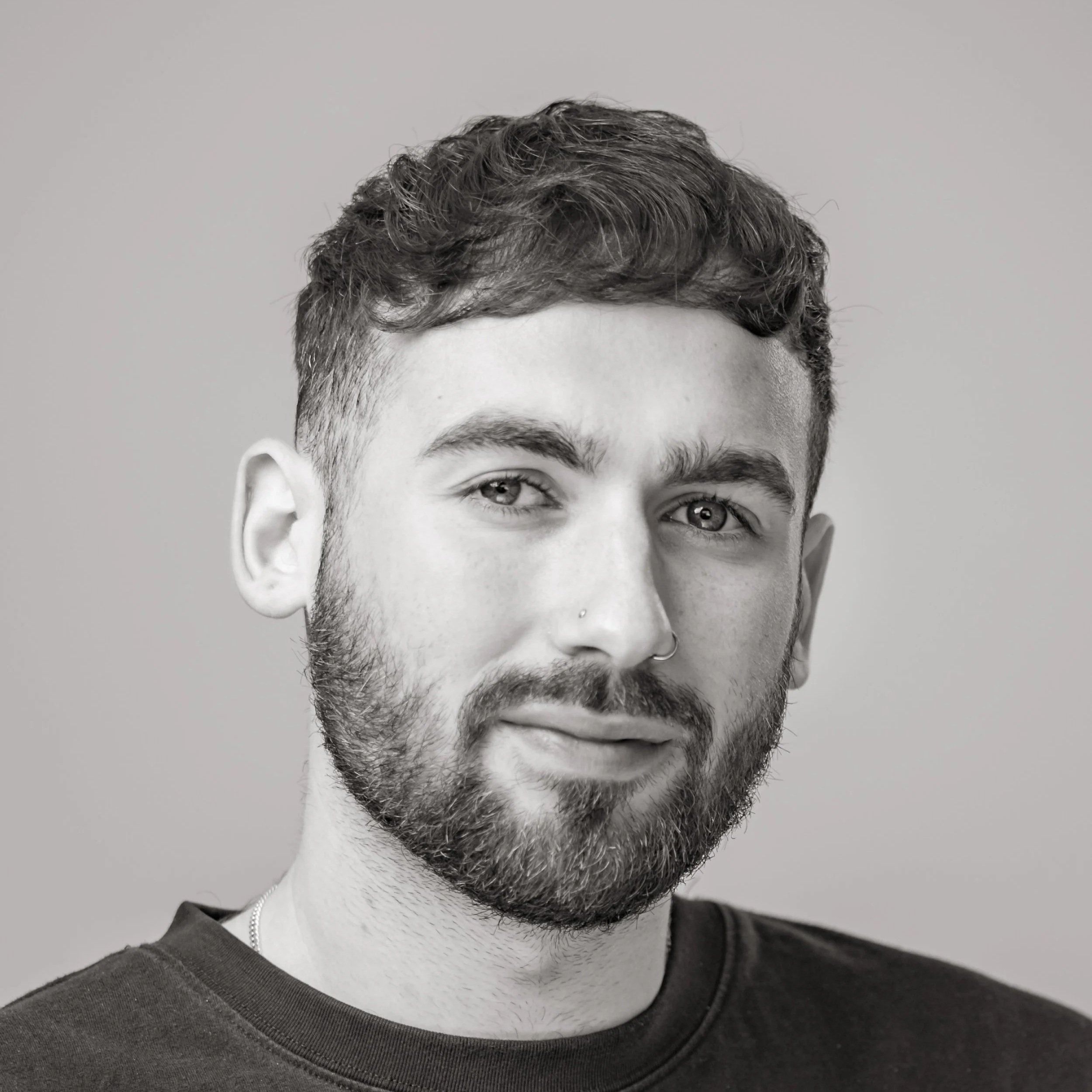 Black and white portrait of a young man with short curly hair, beard, and a nose ring, wearing a dark shirt against a plain background.