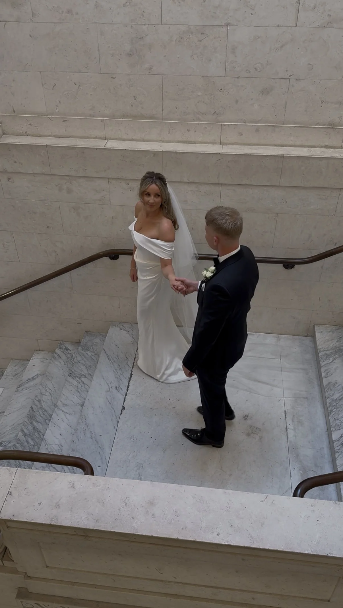 A bride and groom holding hands on a staircase in a building with beige stone walls, celebrating their wedding.