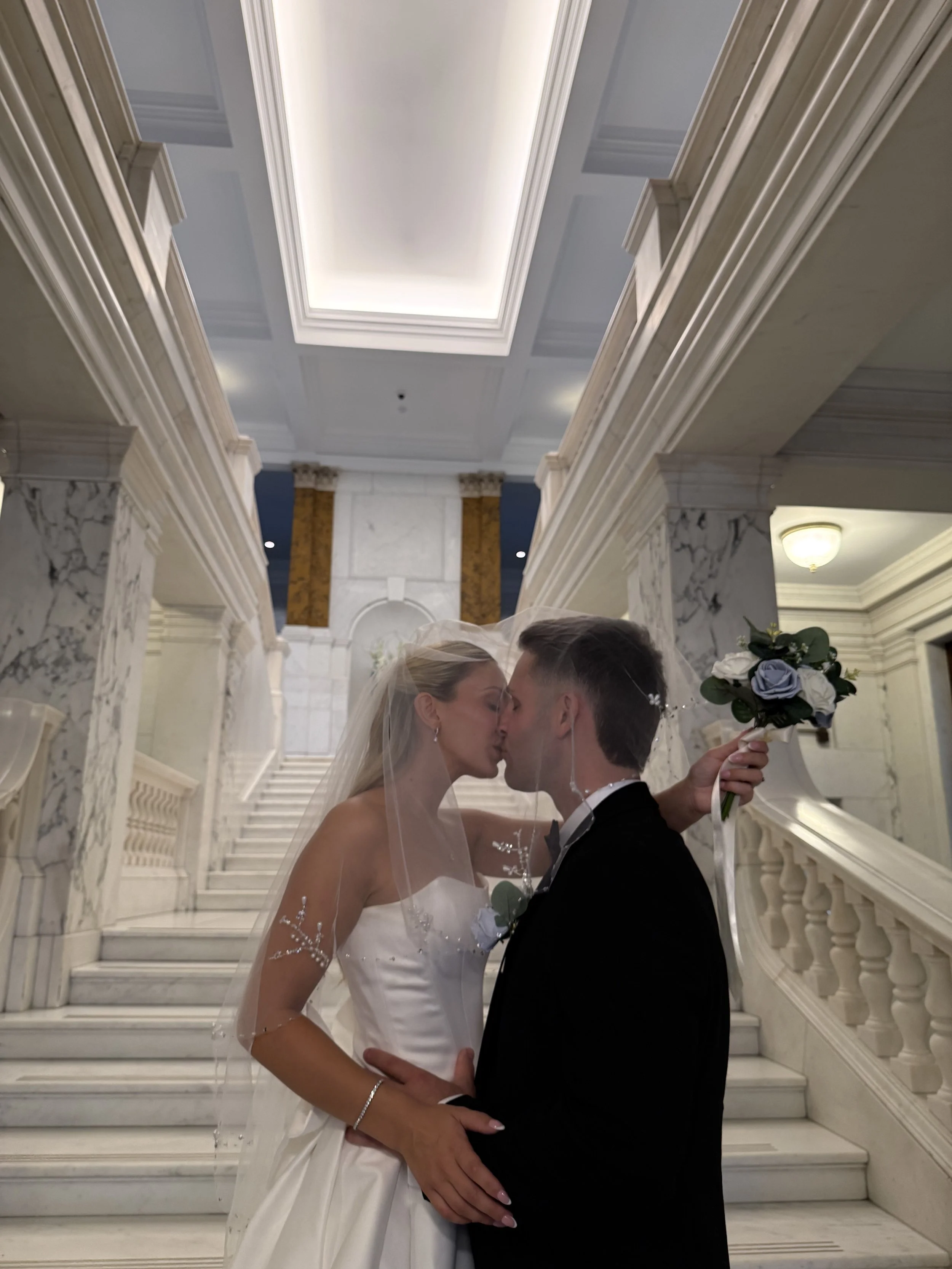 A newly married couple share a kiss on town hall stairwell, the bride holding her bouquet as they celebrate just after the ceremony.