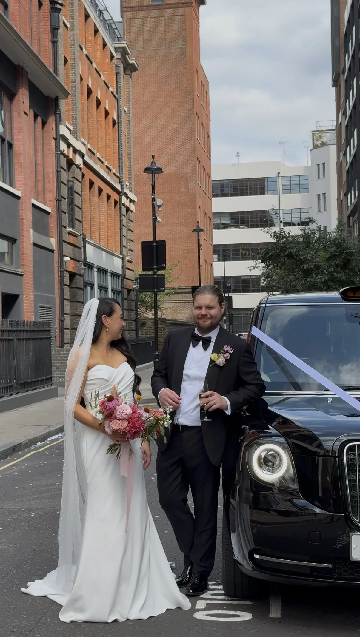 Bride and groom standing next to a black luxury car decorated with a white ribbon, smiling and holding drinks, in an urban street with brick and modern buildings.