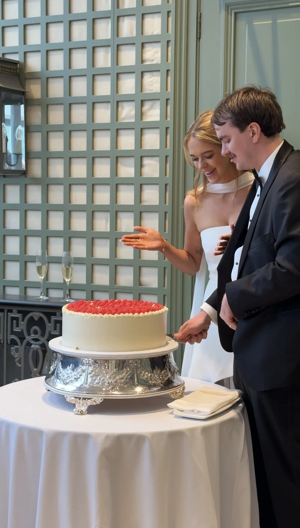 newly married couple cutting their wedding cake, with the groom slicing the cake and the bride smiling happily beside him.